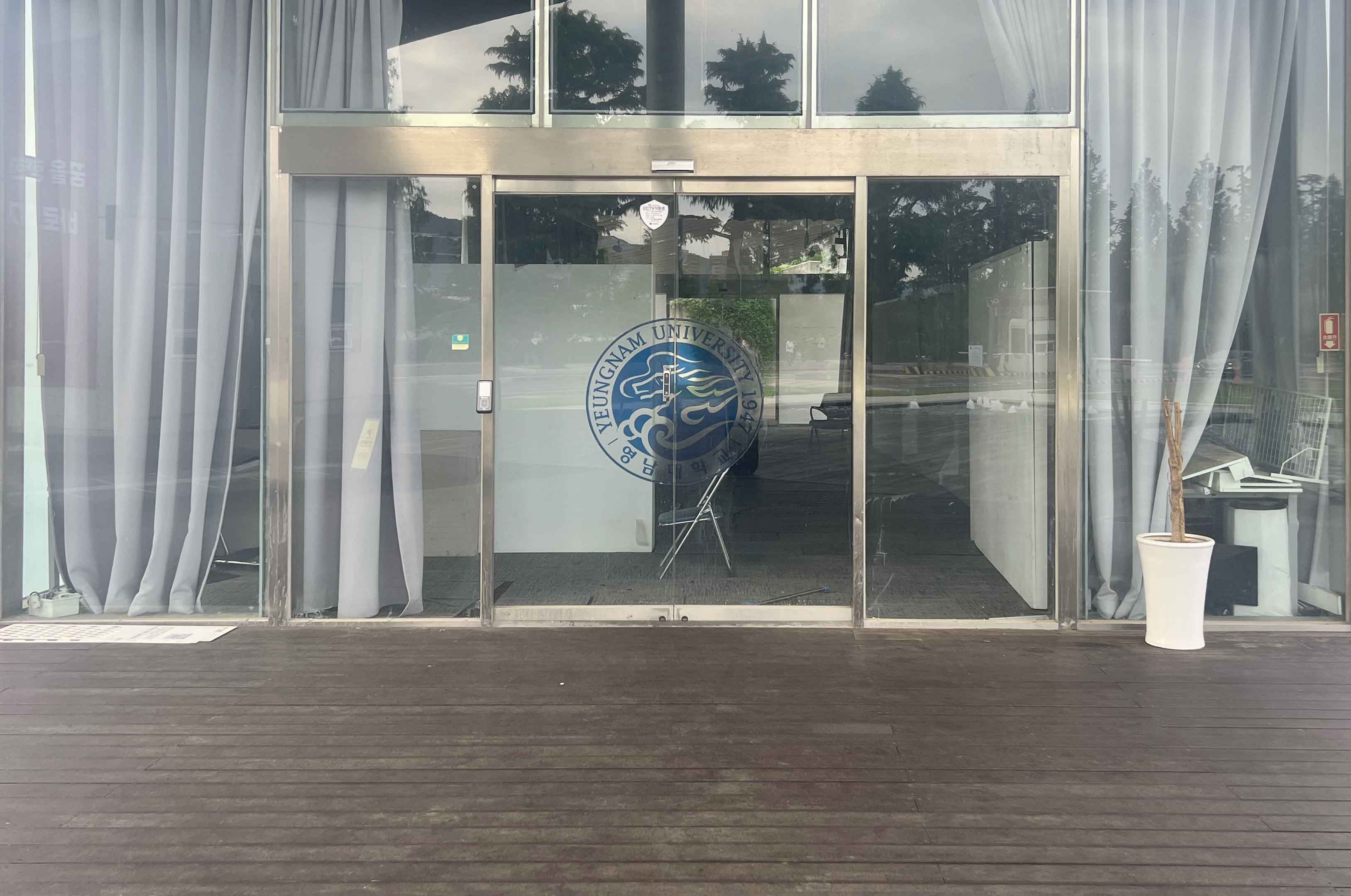 통유리창 너머로 양쪽 끝에 하늘색 커튼이 보인다. 중간에는 영남대학교 로고가 그려진 유리문이 있다. 바닥은 어두운 갈색 나무판으로 이루어져 있다.