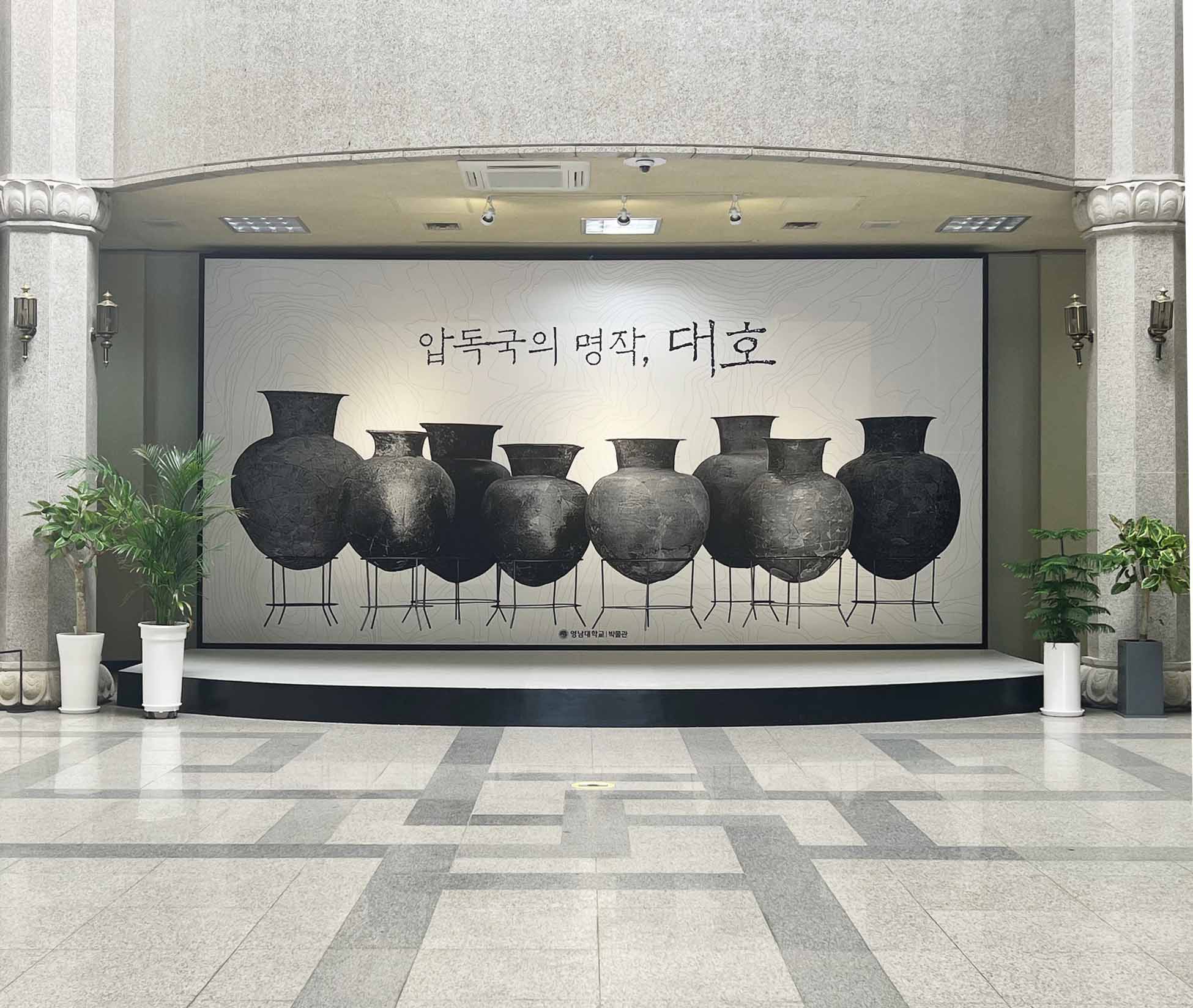 양쪽 끝에 밝은 기둥, 화분 2개가 대칭으로 있다. 중간에는 여러 도자기가 그려진 사진이 있다. 바닥은 밝은 회색 바탕에 어두운 회색으로 네모 모양과 비슷한 기하학적인 무늬가 있다.