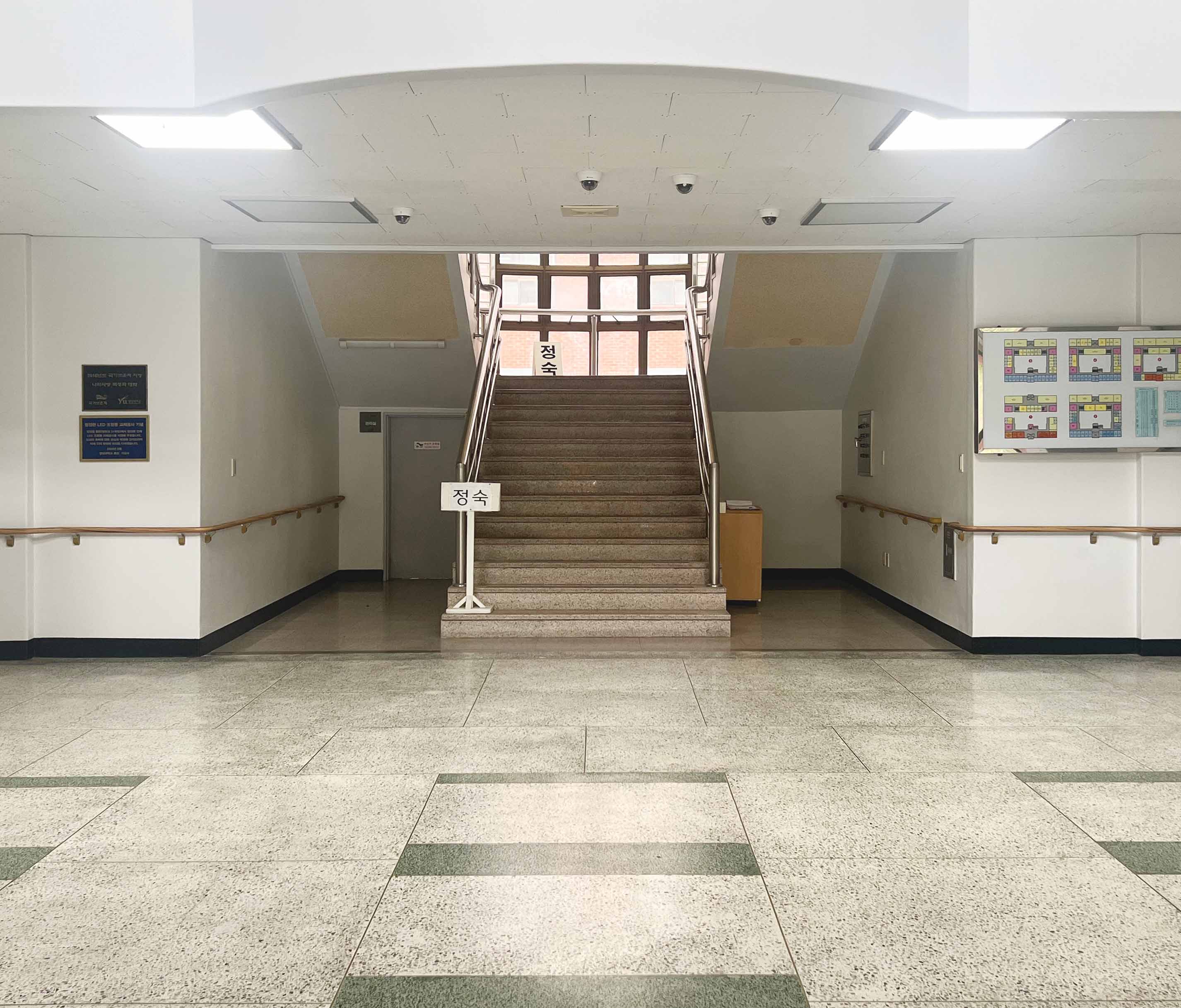 천장에 2개의 힌색 조명이 있다. 중앙엔 계단이 정면으로 보이고 양옆으로 위쪽으로 올라가는 계단이 있다. 벽과 천장은 흰색, 계단은 밝은 갈색, 바닥은 밝은 회갈색 바탕에 부분적으로 가로로 긴 초록색 타일로 장식되어 있다.