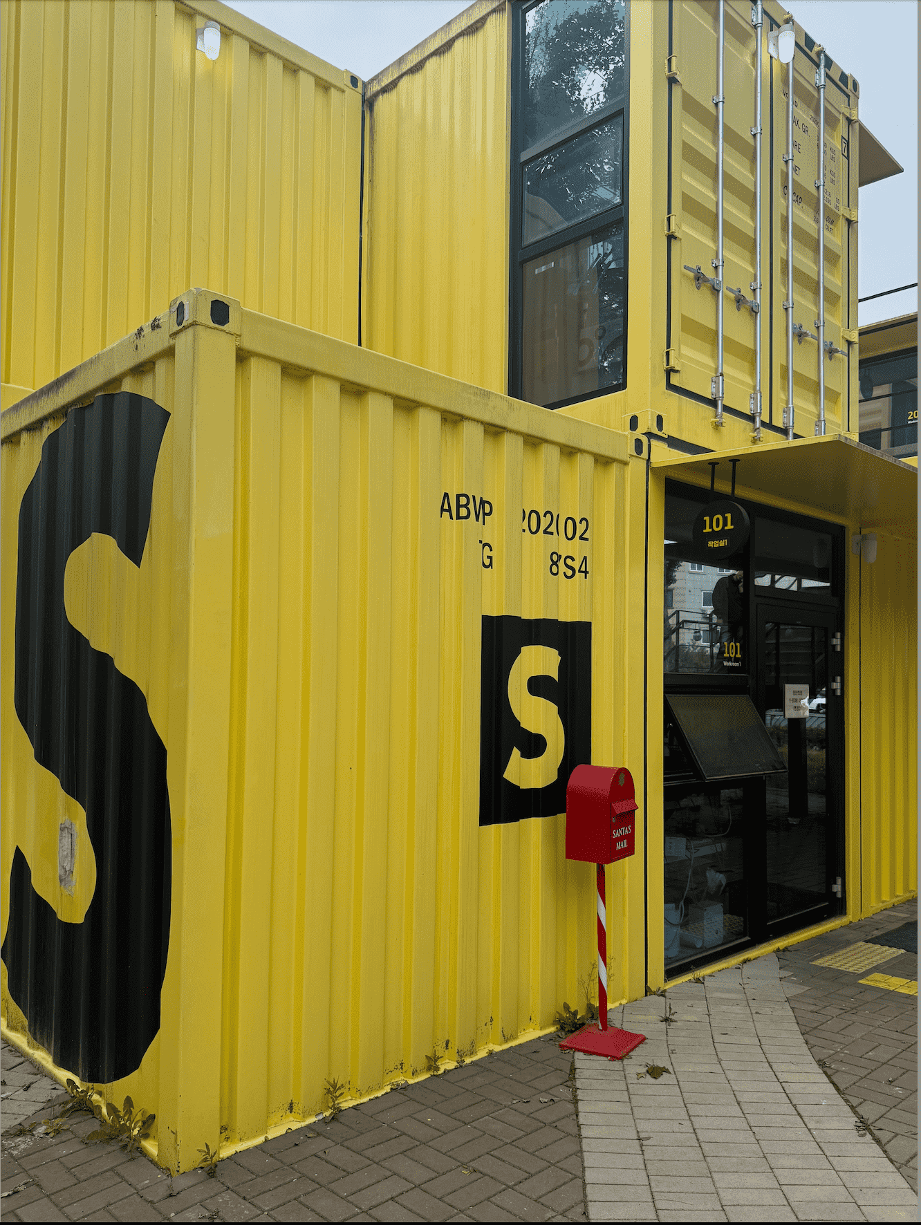 노란 컨테이너로 만들어진 건물의 벽을 찍은 것이다.
                벽의 왼쪽에 서서 오른쪽 방향을 찍었다.벽에는 작게 'ABVP 202002 HG 87S4'라고 크게 'S'가 적혀있다.건물 앞에는 빨간 우체통이 하나 있다.