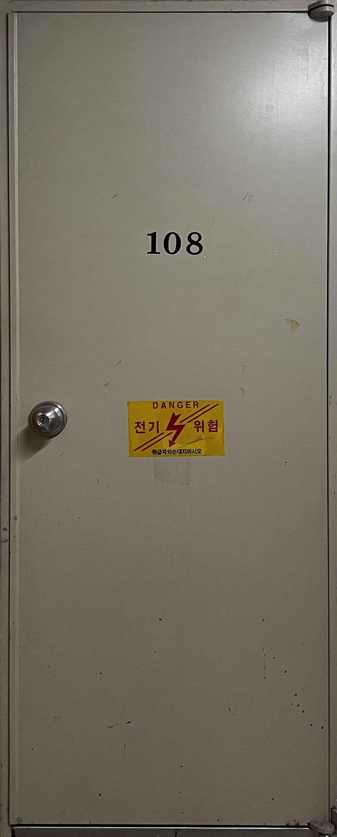 노란빛이 조금 더 도는 베이지색의 철제문이다. '108'이 새겨져있고 손잡이 옆에 노란색 바탕에'전기 위험'이라는 빨간 글씨가 적힌 스티커가 붙어있다.