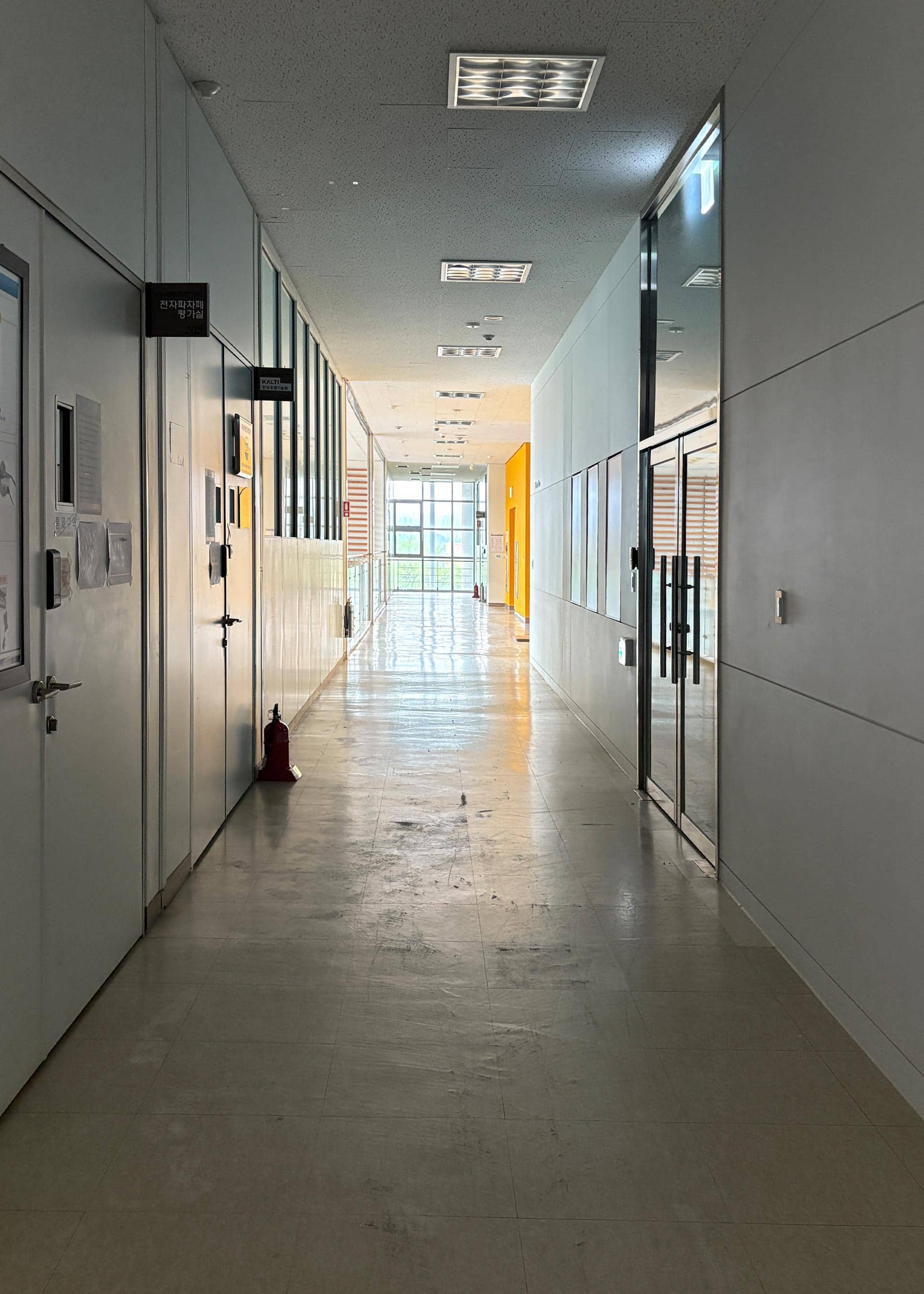 영남대학교 산학협력단 2층 중복도. 왼쪽 벽에는 평가실들이 있고 문 옆에는 빨간 소화기가 있다. 오른쪽 벽에는 유리로 된 문이 하나 있다. 복도 끝에는 빛이 들어오는 큰 창문이 있다. 