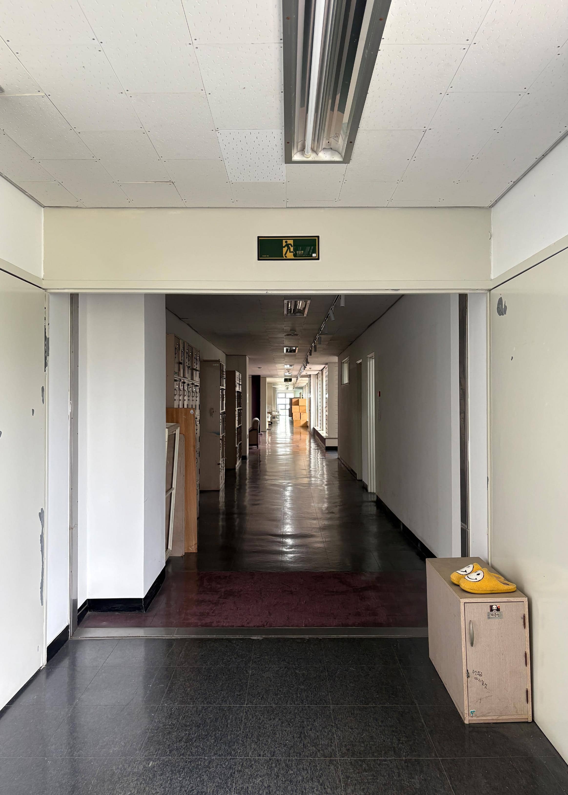 영남대학교 건축관 3층 중복도. 오른쪽엔 작은 사물함과 그 위에 미소를 띤 흰색 얼굴이 그려져 있는 노란 슬리퍼가 있다. 왼쪽 벽에는 사물함으로 나열되어 있다. 