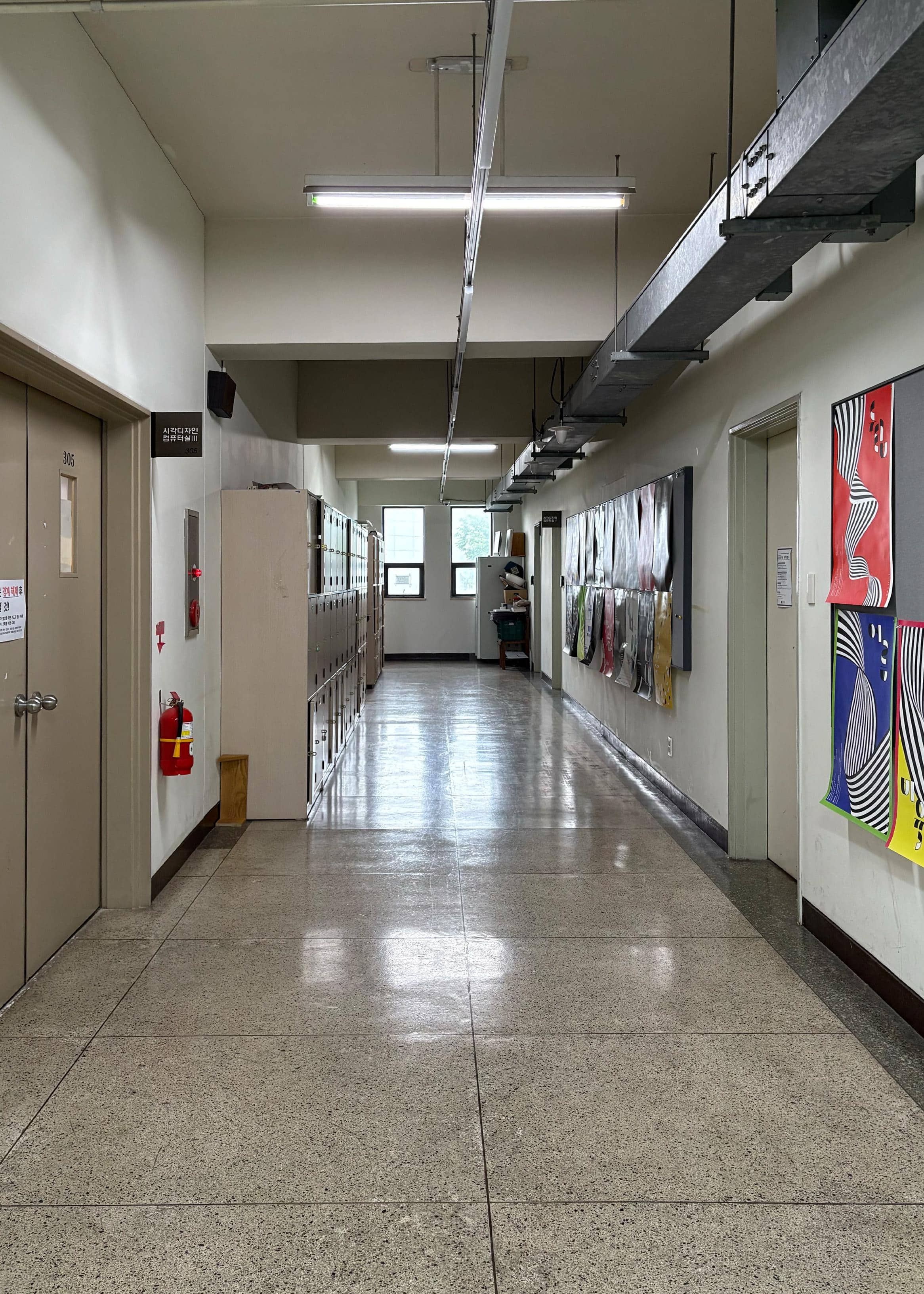 영남대학교 디자인관 3층 중복도. 왼쪽 벽에는 시각디자인과 컴퓨터실이 있고 옆에는 세로로 긴 사물함들이 여러 개 있다. 오른쪽 벽에는 여러 포스터와 강의실들이 있다. 복도 끝에는 세로로 긴 창문이 두 개 있다.