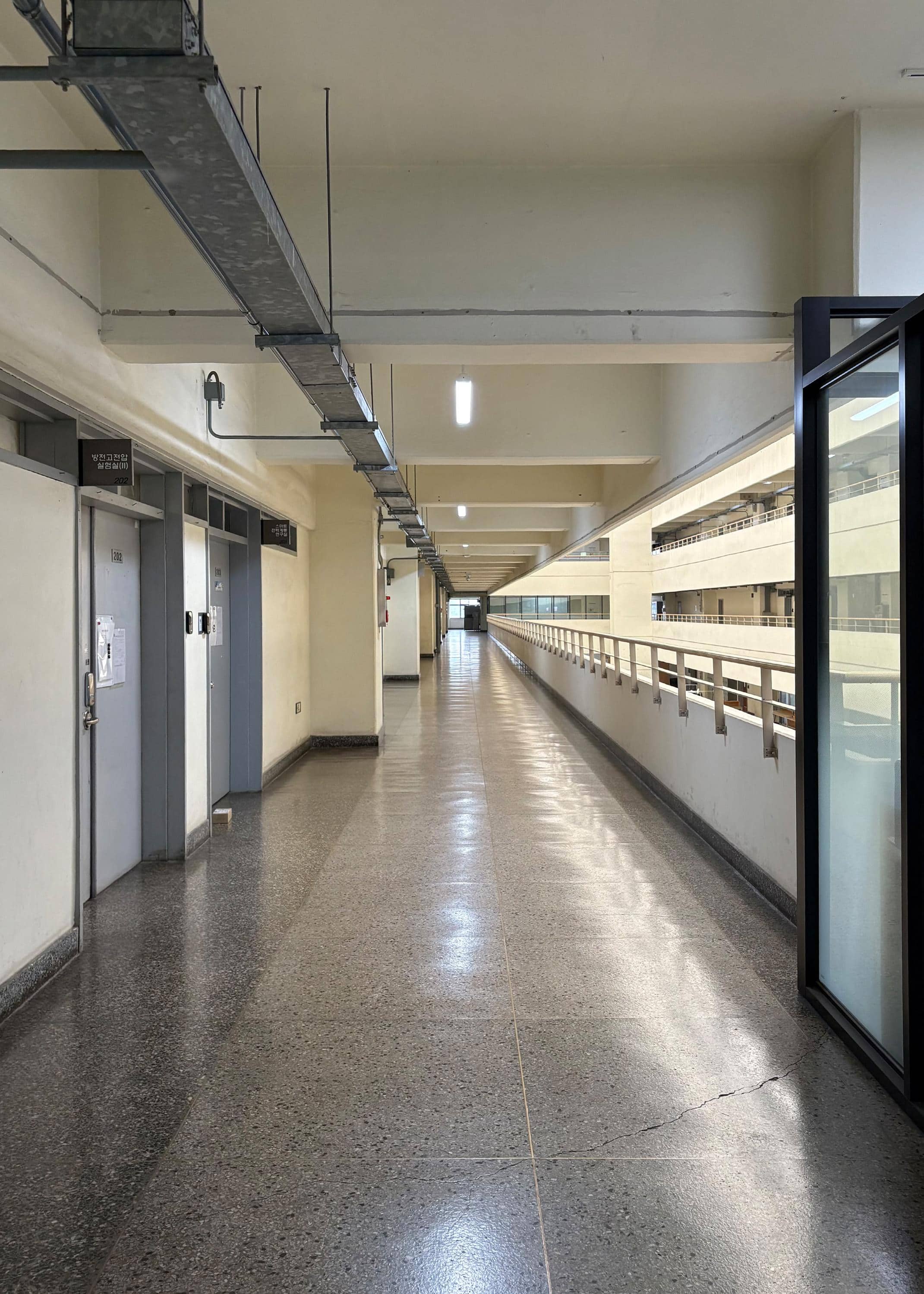 영남대학교 기계관 2층 편복도. 회색 철문이 두 개 있고 흰색 기둥이 반복적으로 있다.  오른쪽에는 유리로 이루어진 자습실 벽이 보이고 그 옆엔 난간이 복도 끝까지 있다. 복도 끝에는 창문이 하나 있다.