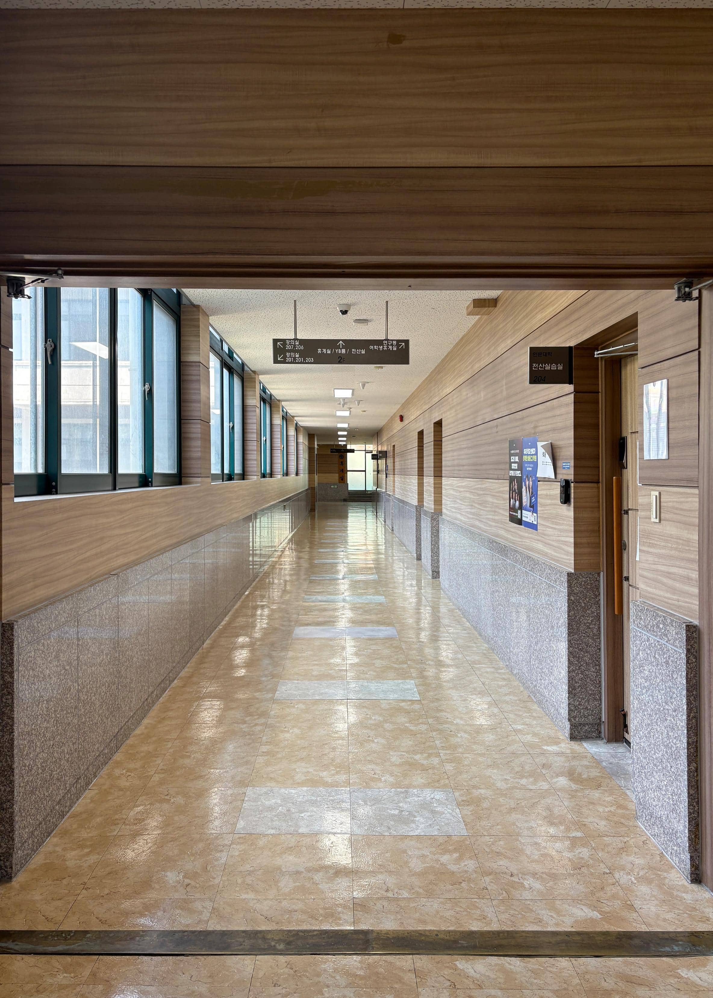 영남대학교 인문관 2층 편복도. 왼쪽 벽에는  초록색 창틀인 창문이 반복적으로 있다. 오른쪽 벽에는 실습실이 세 개 있다. 복도 끝에는 다른 강의실과 이어져 있는 유리문이 있다.