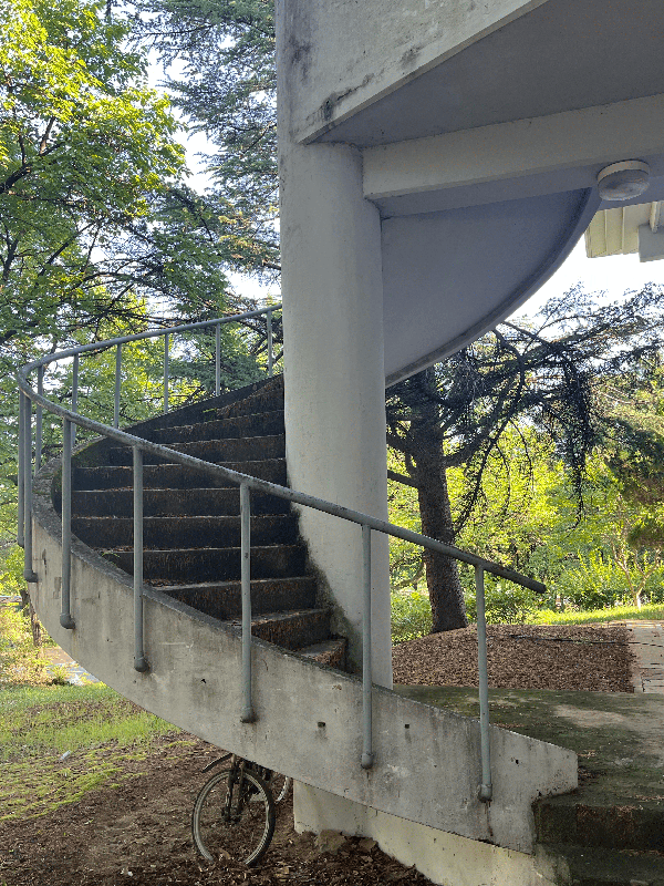 흰색 기둥을 가준으로 돌아가는 탁한 갈색 타일의 나선형 게단을 옆에서 찍은 사진. 계단 뒤에는 나무와 풀이 보인다.