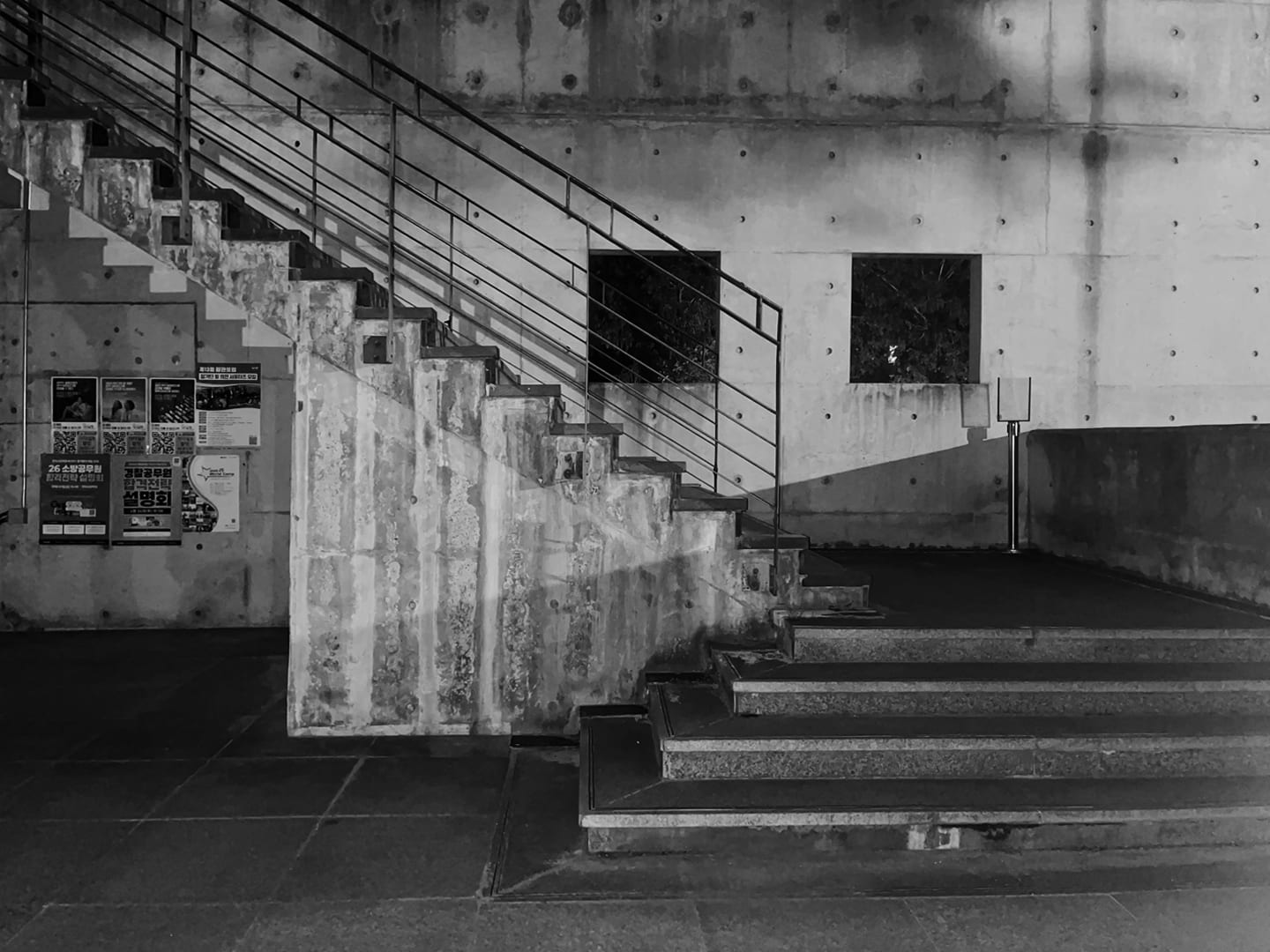 회색 콘크리트 벽 앞에 있는 콘크리트 계단이 보이는 흑백사진. 왼쪽 계단은 위로 비스듬히 뻗어 있고 검은색 금속 난간이 설치되어 있다.