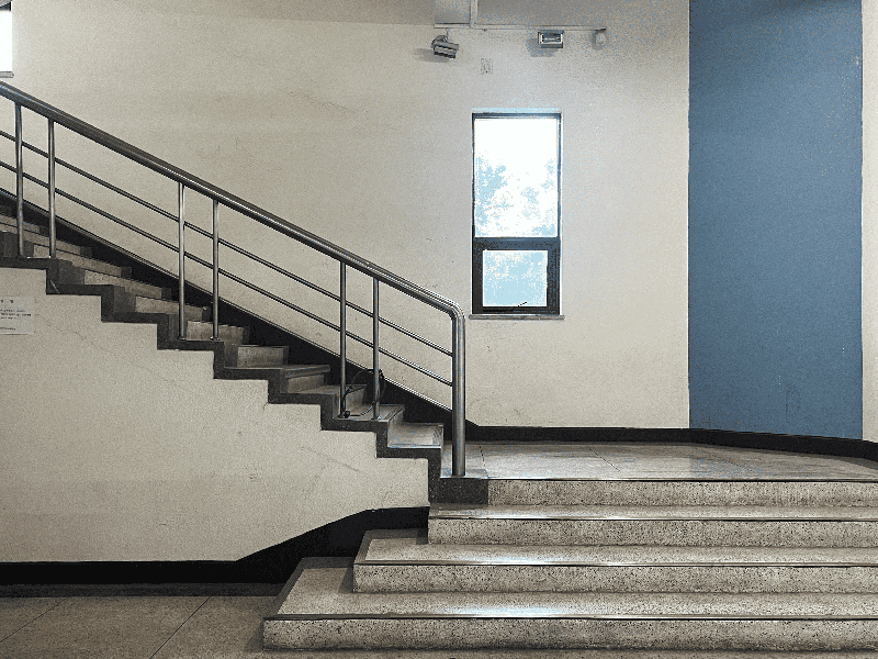 실내 계단이 보이는 사진. 왼쪽에는 은색 금속 난간이 설치된 회색 계단이 위로 비스듬히 뻗어 있고, 오른쪽에는 아래에서 위로 연결되는 또 다른 회색 계단이 보인다. 계단 중간 벽에는 창문이 있고, 창문 오른편 벽은 파란색으로 칠해져 있다.