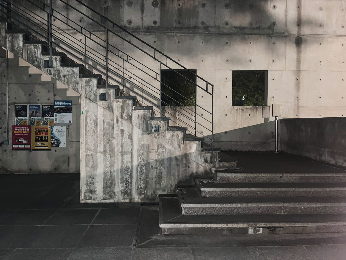 회색 콘크리트 벽 앞에 있는 콘크리트 계단이 보이는 사진. 왼쪽 계단은 위로 비스듬히 뻗어 있고 검은색 금속 난간이 설치되어 있다. 계단 중간쯤에 창문 두 개가 나란히 있으며, 오른쪽에는 아래에서 위로 연결되는 또 다른 콘크리트 계단이 보인다.