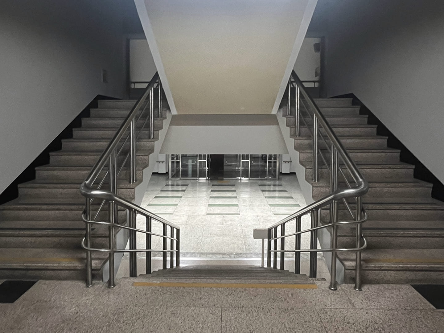 위로 뻗은 계단 두 개가 양쪽으로 뻗어있는 사진. 계단을 내려가면 건물 밖으로 나갈 수 있는 유리문이 보인다.