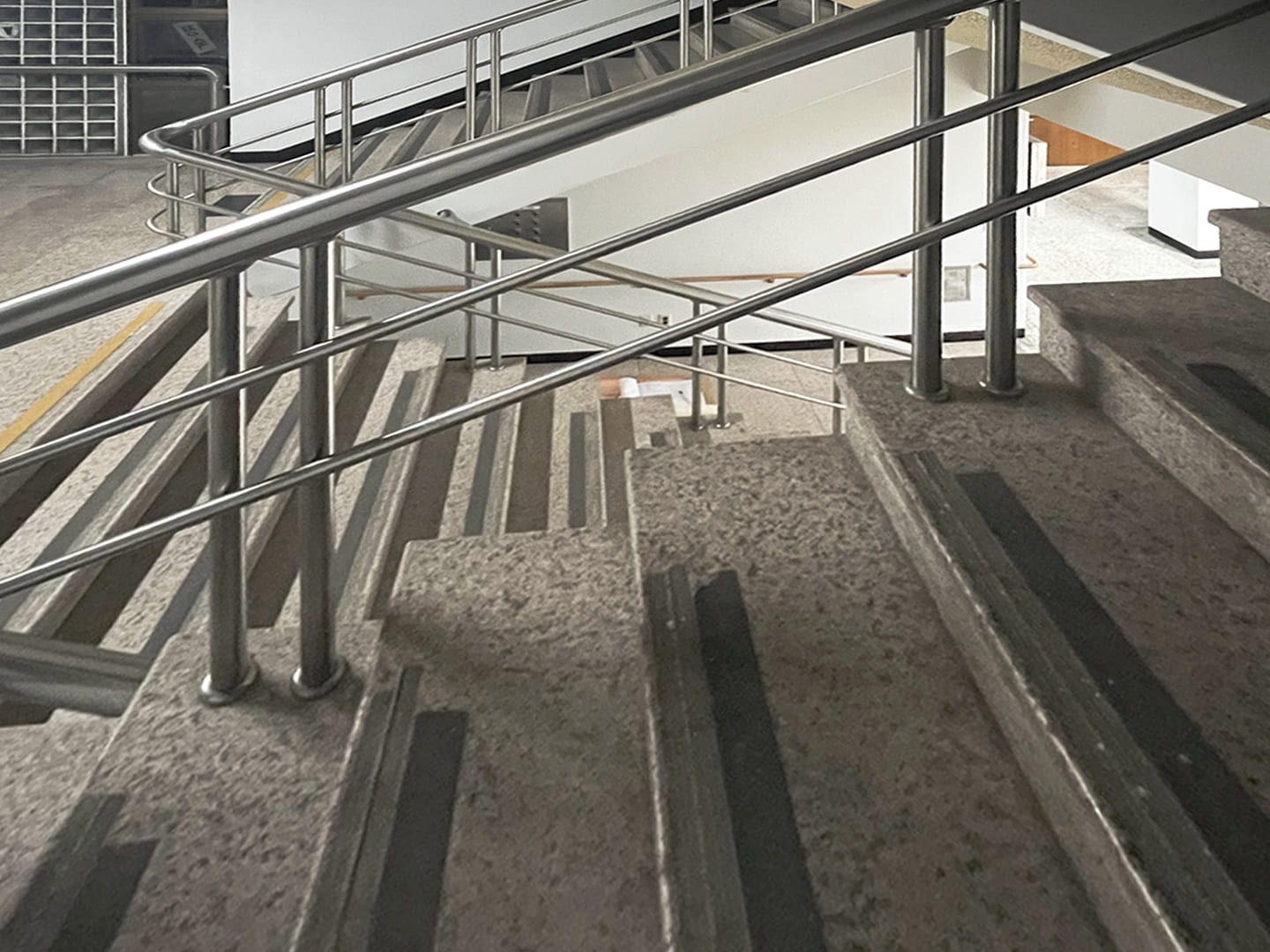 계단 난간과 계단이 보이는 실내 풍경 사진. 왼쪽에는 은색 금속 난간이 위로 비스듬히 뻗어 있고, 오른편에는 회색 계단이 아래로 이어진다. 계단에는 검은색 미끄럼 방지 패드가 일정한 간격으로 부착되어 있다.