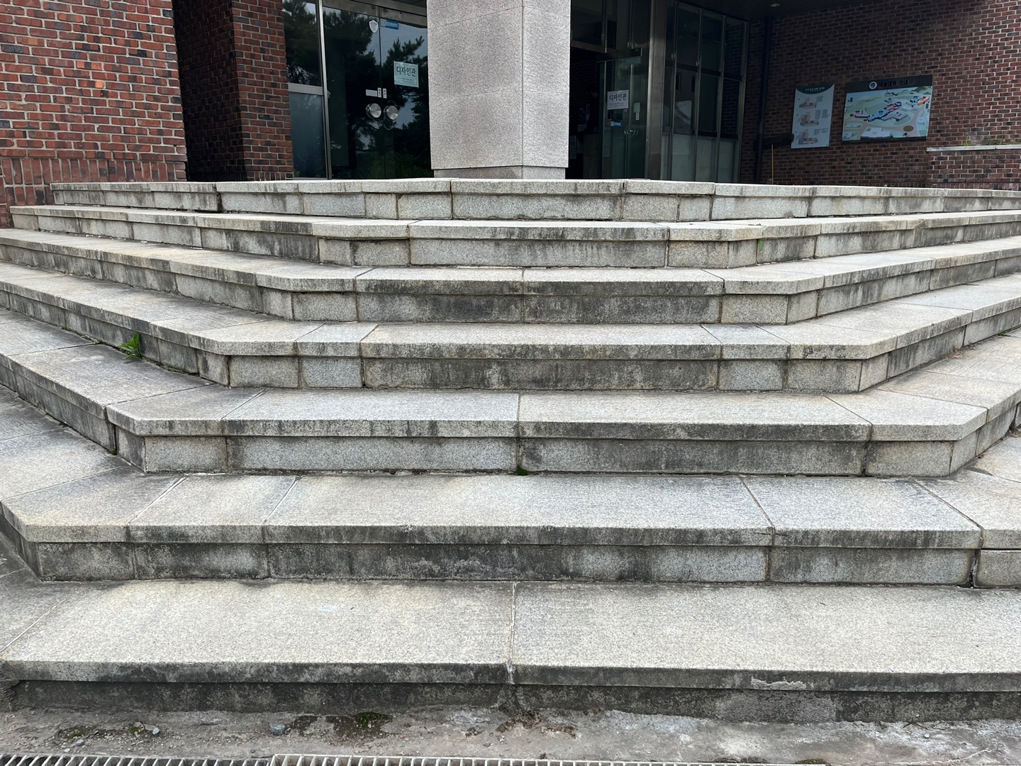 건물 입구에 있는 넓고 낮은 흰색 타일의 콘크리트 직선 계단의 이미지. 계단 위에는 유리문,기둥 등이 보인다.