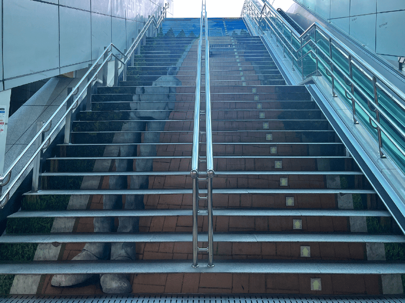 넓고 긴 계단이 정면으로 보이는 사진. 계단 가운데에는 은색 금속 난간이 위로 길게 뻗어 있으며, 계단 전체에는 벽돌길 위에 서 있는 사람의 다리가 그려진 착시 미술 작품이 그려져 있다. 계단 오른편으로는 에스컬레이터가 보인다.
