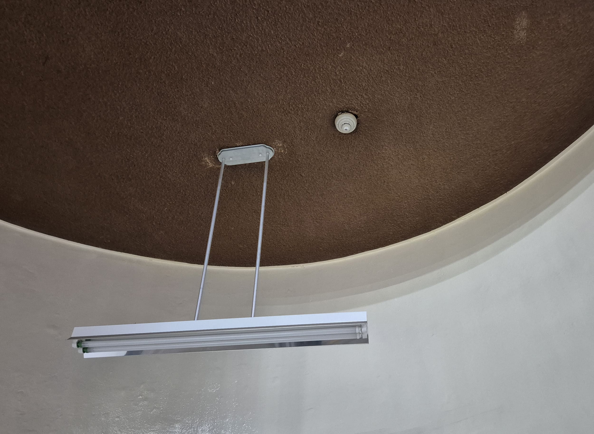 옆쪽에서 찍은 동그란 형태의 천장 사진이다. 천장의 색상은 갈색이고, 벽면의 색상은 아이보리 색이다. 철제 와이어로 매달린 직사각형의 LED조명이 천장의 중심에서 수직에서 내려오는 형태를 하고 있다.