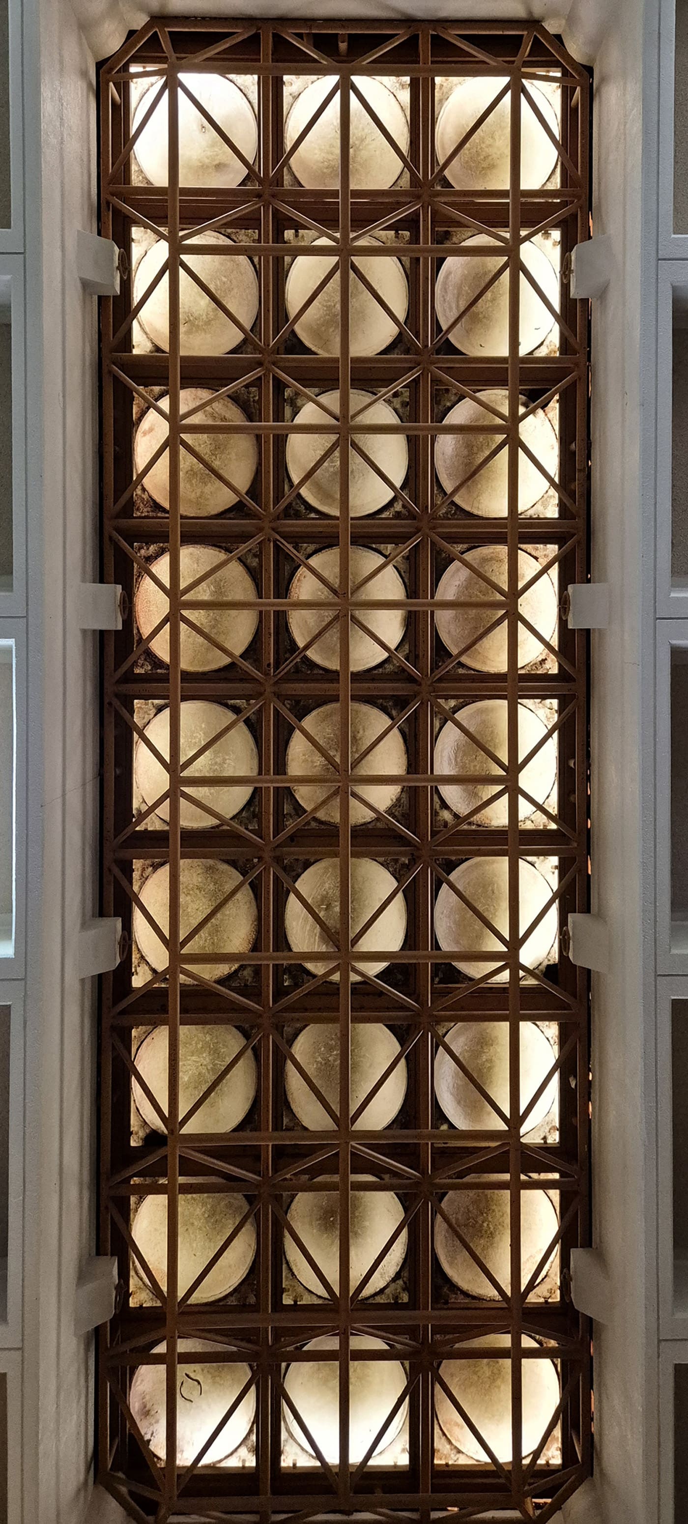 정사각형 격자 구조 안에 원형의 조명이 놓여져 있는 구조가 반복적으로 배치되어 있는 천장 사진이다. 천장의 격자 부분은 짙은 갈색, 안쪽 조명은 연한 아이보리색이다. 천장을 정면에서 찍은 세로로 긴 사진이다.