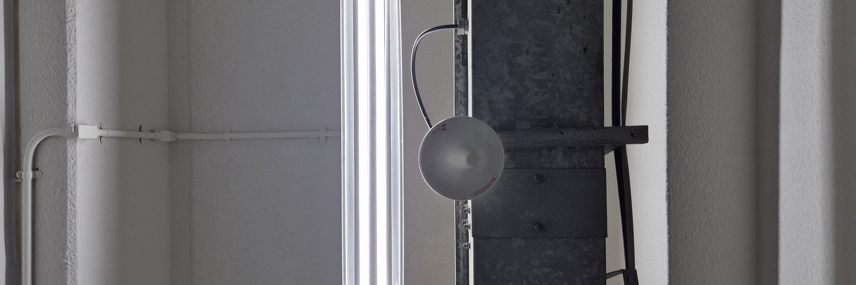 흰색의 천장과 천장 가운데에 세로로 나란히 배치된 긴 형광등이 있다. 사진의 가운데로부터 살짝 오른쪽에는 어두운 회색빛의 넓은 금속 기둥이 보인다.천장을 정면에서 찍은 가로로 긴 사진이다.