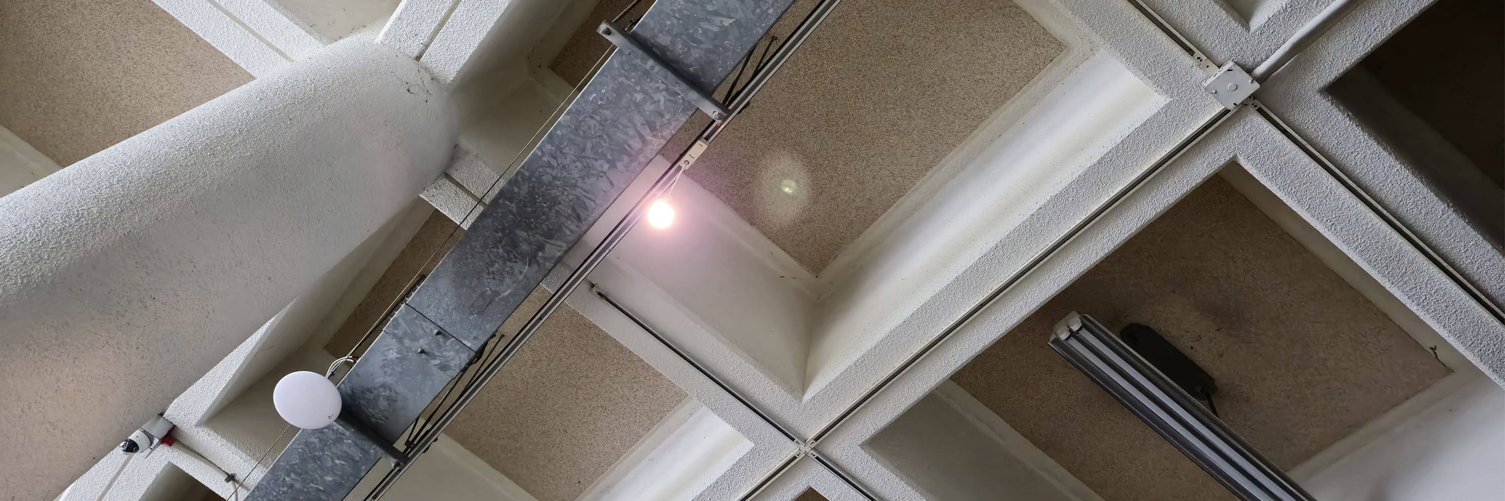 사각형 격자 형태로 나열된 구조의 천장 사진이다. 천장의 격자 부분은 아이보리색이고, 격자 안쪽의 천장 색은 옅은 황갈색 빛을 띤다. 왼쪽에 세로로 긴 기둥이 있다. 중앙에서 왼쪽 부분에는 밝게 빛나는 작은 조명이 설치되어 있다. 천장을 측면에서 찍은 가로로 긴 사진이다.