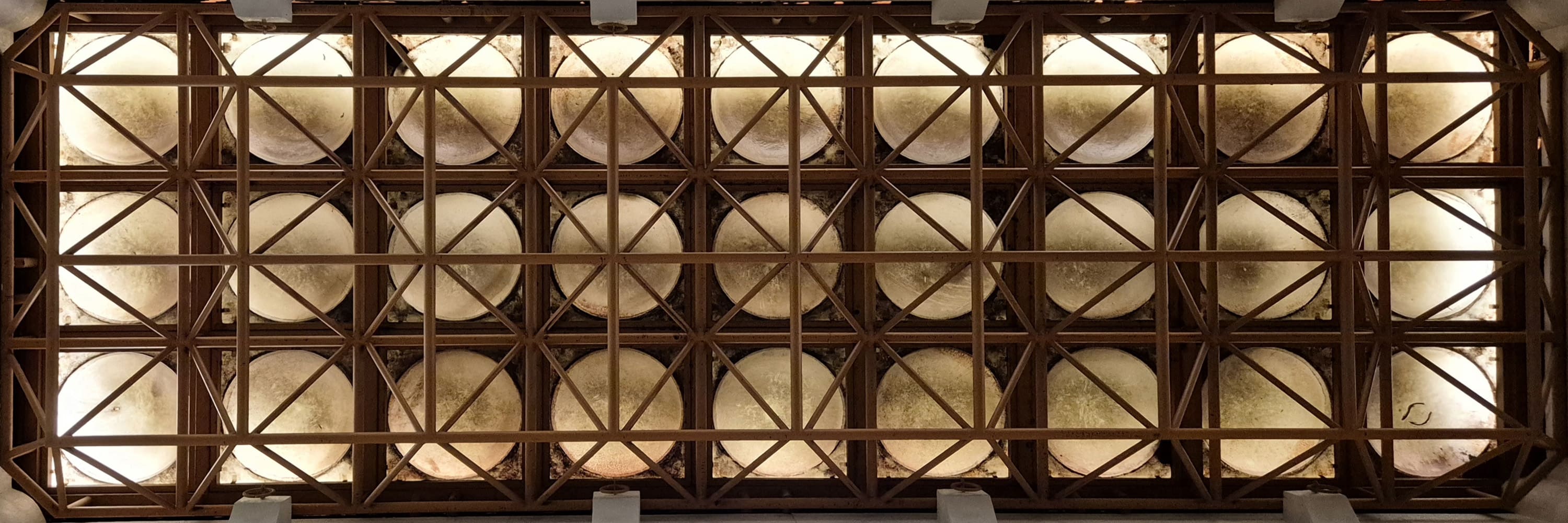 정사각형 격자 구조 안에 원형의 조명이 놓여져 있는 구조가 반복적으로 배치되어 있는 천장 사진이다. 천장의 격자 부분은 짙은 갈색, 안쪽 조명은 연한 아이보리색이다. 천장을 정면에서 찍은 가로로 긴 사진이다. 