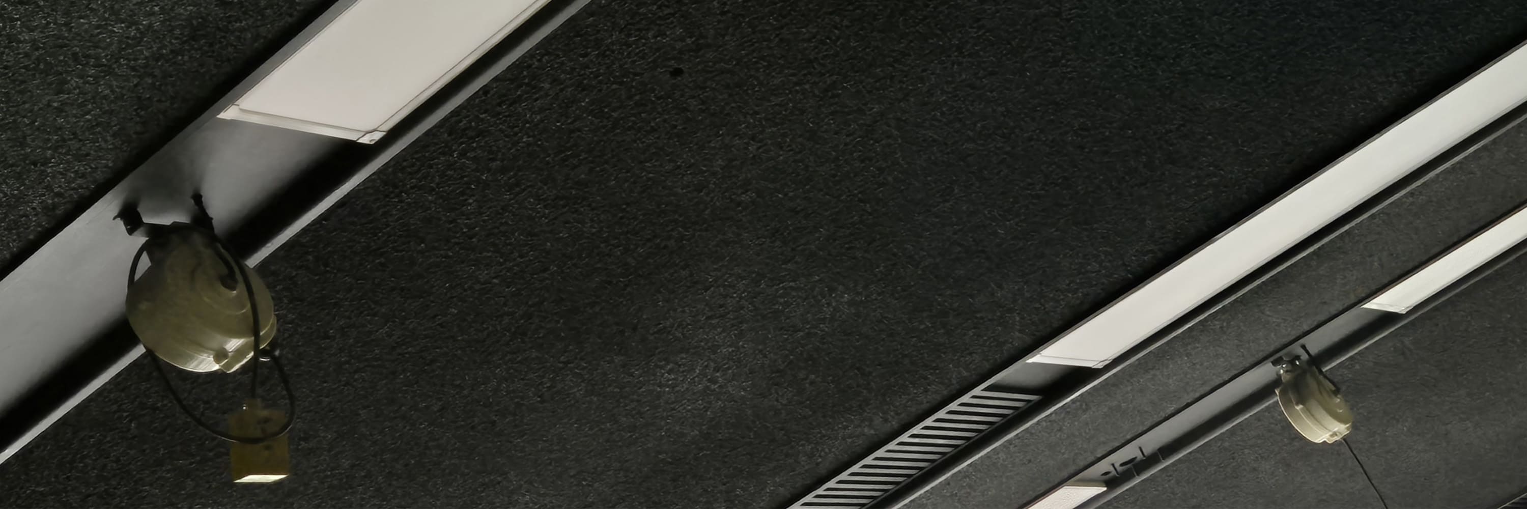 검은색 배경의 천장 사이 길쭉한 조명이 규칙적으로 배치된 형태의 천장 사진이다. 천장의 표면은 약간 까끌한 질감처럼 보인다 천장의 조명이 비스듬하게 찍힌 사진이다.