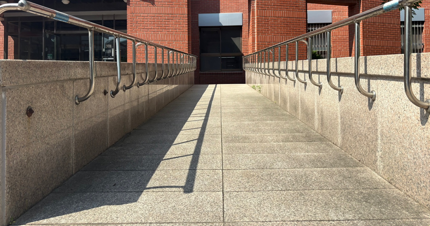 붉은 벽돌 건물 앞에 금속 스테인리스 손잡이와 직사각형의 타일로 이루어진 직선형의 미색 램프를 정면,손잡이 높이에서 본 사진 입니다.