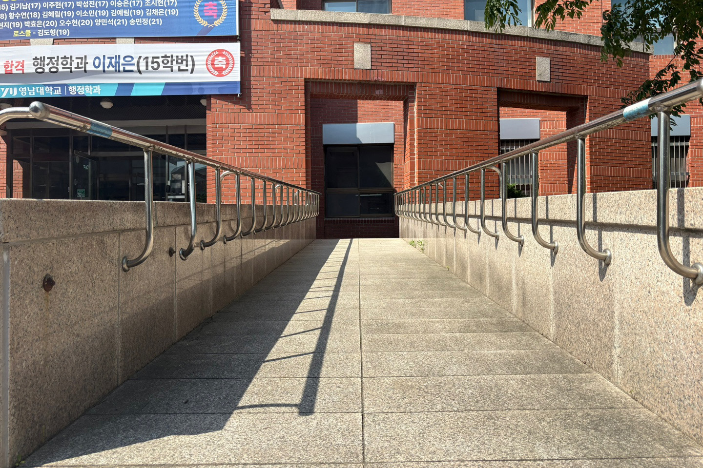 뒤쪽의 둥글고 붉은 벽돌의 건물과 이어지는 스테인리스 손잡이의 미색 직선형 램프를 정면에서 찍은 사진 입니다.