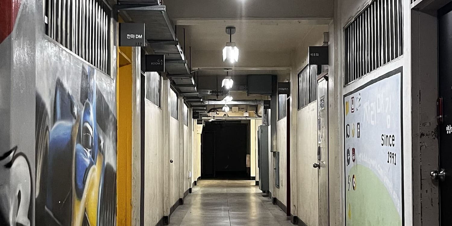 중간에 소실점이 있는 사진을 세로로 3분할 한 사진의 중간. 오른쪽 벽에는 검정색 문, '자리매김 1991'이 적힌 하늘색과 초록색,여러개의 아이콘의 벽화와 세로형 철제 창살이 빼곡히 있는 가로로 긴 직사각형 창문, 옅은 회색의 철제 문과 사우회의 안내판, 같은 창문, 갈색 문, 창문 순으로 있으며 끝에 철제 박스가 있다. 왼쪽엔 회색 벽에 파란색, 노란색이 섞인 레이싱카의 벽화가 있다. 그 위에는 쇠창살이 있는 창문, 옆에는 노란색 문틀과 '천마DM'의 안내판, 창문 두 개가 더 있다. 왼쪽 위에는 철제 덕트가 설치되어있다.천장에는 노출형 2구형광등. 왼쪽 형광등은 빠져있다.바닥은 테라조 타일 2번 사진의 중간