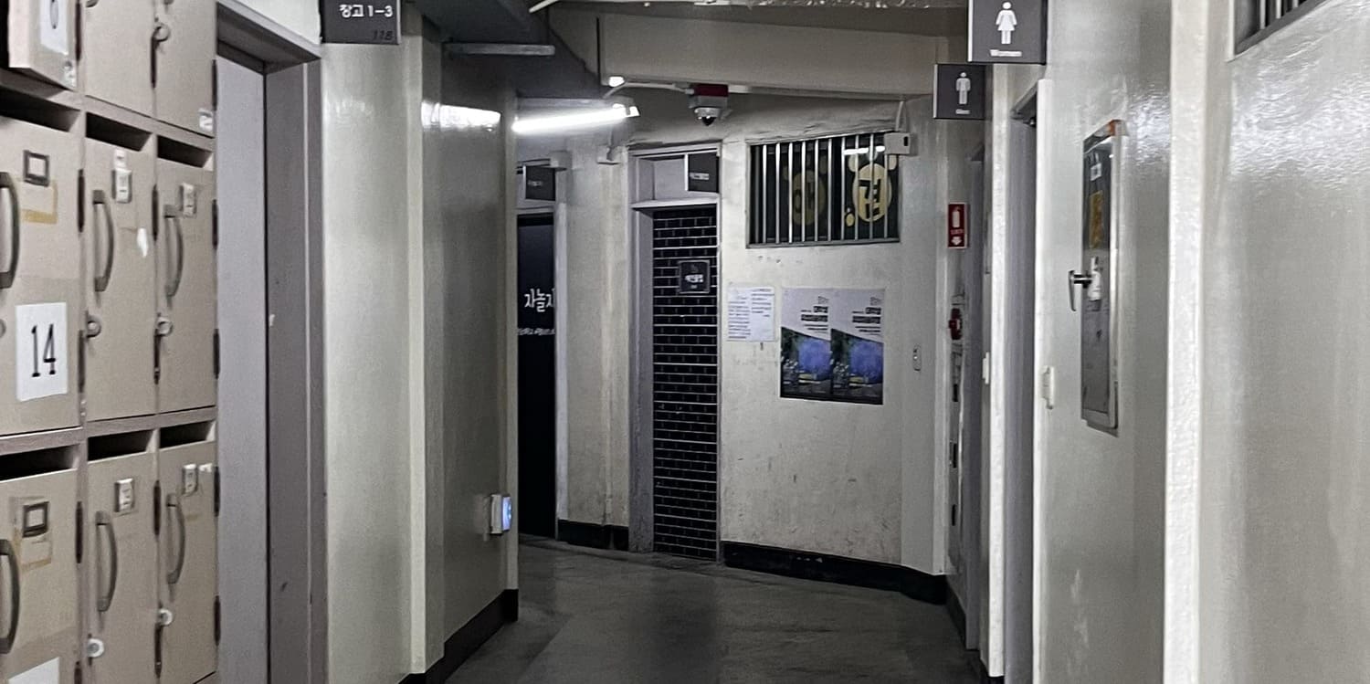 중간에 소실점이 있는 사진을 세로로 3분할 한 사진의 중간.끝에서 왼쪽으로 복도가 꺾여있다. 시멘트 벽의 왼쪽에는 벽에 매입된 철제 전기 설비함이 있고, 옆에는 여자,남자 화장실이 있다. 오른쪽 벽에는 사물함과 연보라색빛이 도는 철제 문이 있다. 왼쪽 천장에는 덕트가 이어져있다.3번 사진의 중간.