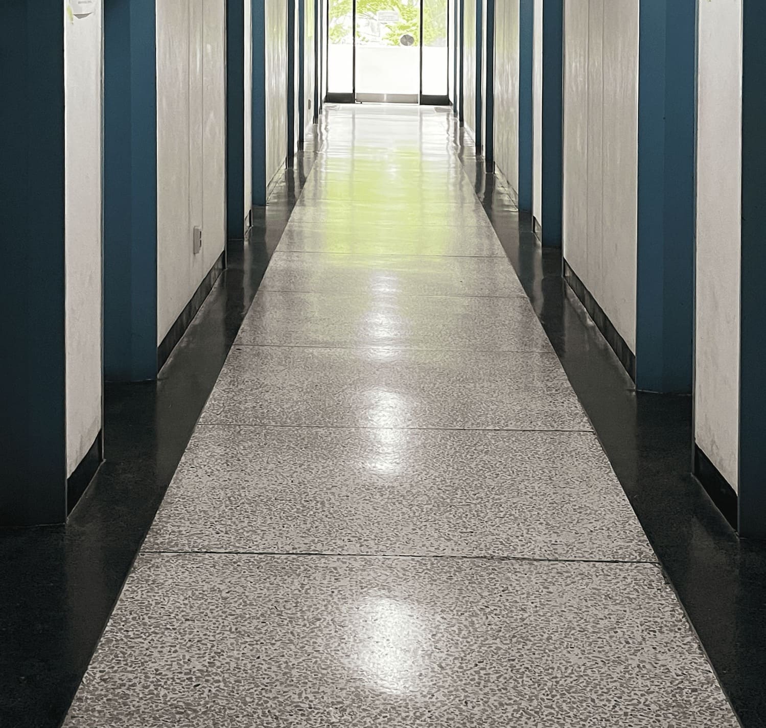 소실점이 중앙에 있는 복도 전체의 중간부터 바닥까지의 사진. 양쪽 벽은 대칭으로 이루어져 각각 7개의 문틀이 보임. 문틀은 모두 짙은 하늘색이다. 복도 끝에 있는 밖으로 나가는 유리문이있다. 바닥엔 큰 테라조 타일이 쭉 나열되어있다. 2번 복도의 아래.
