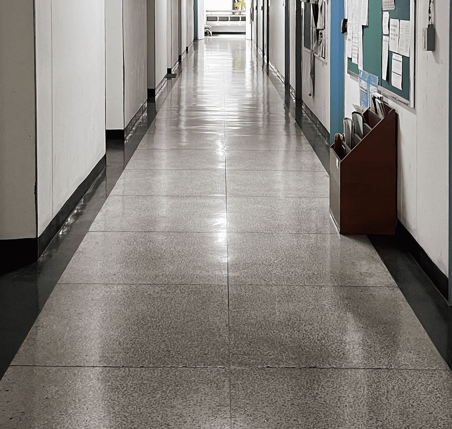 소실점이 중앙에 있는 복도의 중간부터 바닥까지의 사진. 바닥엔 작은 테라조 타일이 2개씩 쭉 나열되어있다. 콘크리트 벽의 왼쪽에는 오목하게 들어간 부분이 규칙적으로 있고, 오른쪽 벽에는 하늘색 문틀이 나열되어있다. 작은 3단 나무 책꽂이도 오른쪽 벽에 붙어 서있다. 그 위에는 게시판이 있다. 3번 복도의 아래