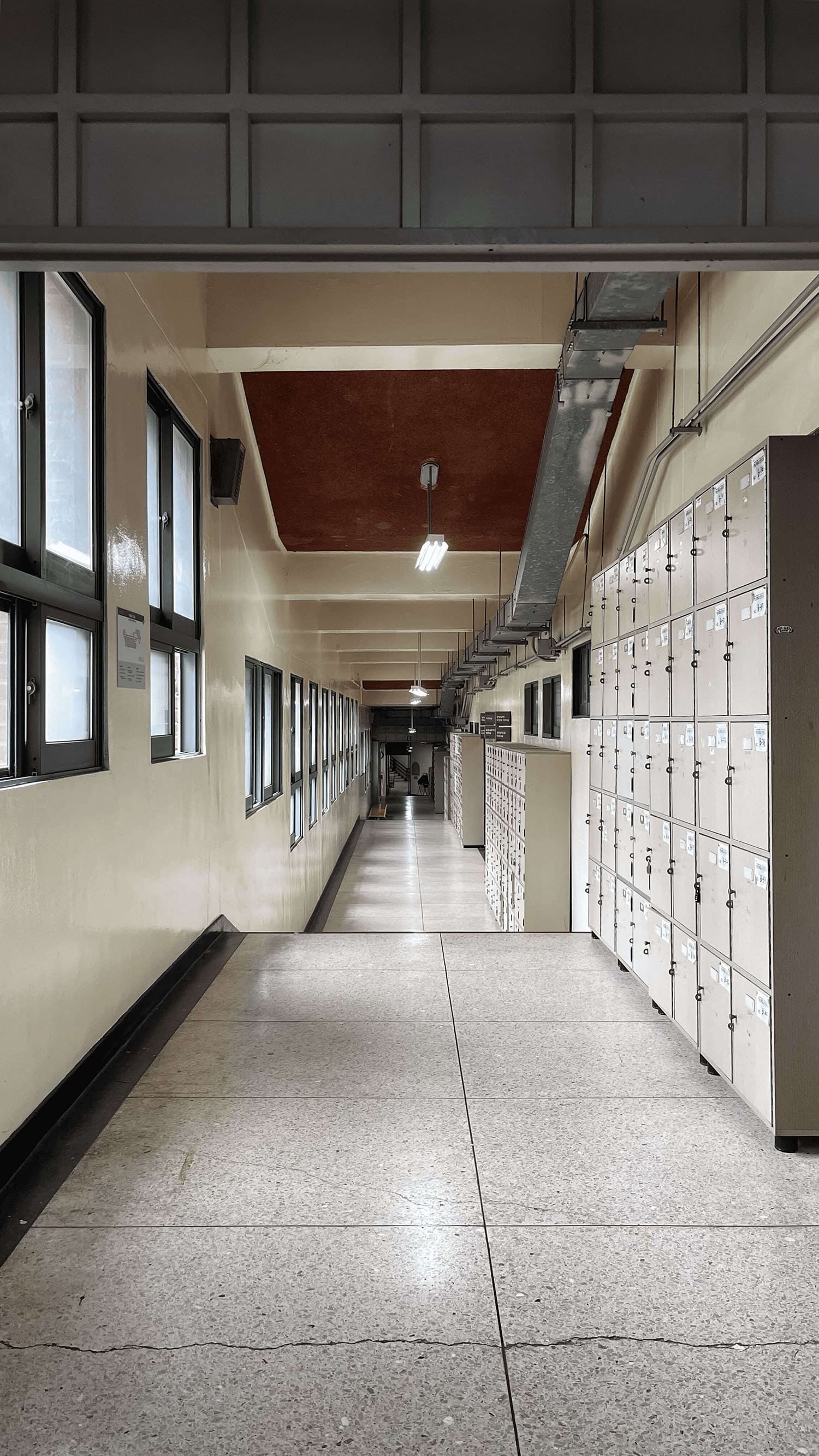 오른쪽 벽면에 사물함이 있는 복도의 사진. 중앙에 소실점이 위치한 채 찍힌 사진.왼쪽 벽면에는 청록색 칠 마감이 된 사각형 창문이 있으며 한 창문이 네 개의 창으로 분할되어 위 두 개는 세로가 긴 직사각형, 아래 두개는 가로가 긴 직사각형 형태이다. 사물함 위 천장에는 철제 덕트도 설치되어있다.바닥은 테라조 타일. 콘크리트 천장엔 규칙적으로 보가 내려와있으며 사진의 가장 윗부분에는 흡음패널이 설치되어있다.