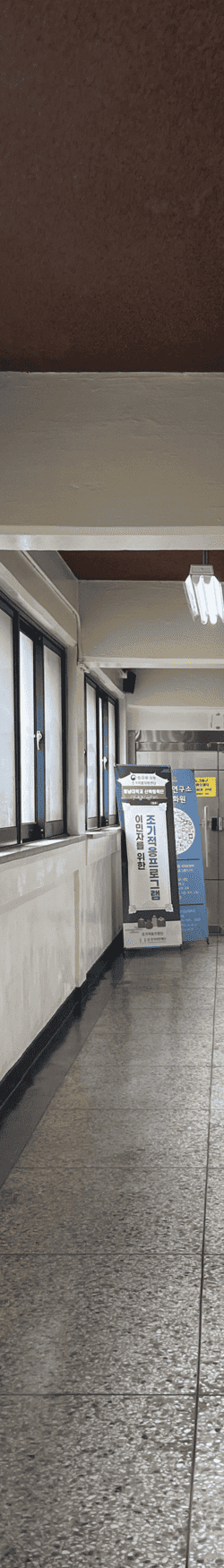 중간에 소실점이 모이는 복도를 세로로 4분할 했을 때의 왼쪽에서 두번째 사진. 콘크리트 천장과 벽. 천장에는 보가 내려와있으며 들어간 부분에는 검붉은색 칠이 되어있고 노출형 형광등이 매달려있다. 복도 끝에는 입간판 2개가 유리문 앞에 있다. 문틀은 철제로 되어있고 바닥은 테라조 타일. 