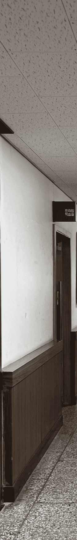중간에 소실점이 모이는 복도를 세로로 4분할 했을 때의 왼쪽에서 첫번째 사진. 왼쪽 벽에는 아래쪽엔 나무몰딩이 되어있고 위쪽엔 프라임 학습실 안내판, 바로 옆에는 나무몰딩된 문틀과 문이 있다.천장은 흡음 텍스 천장이다.