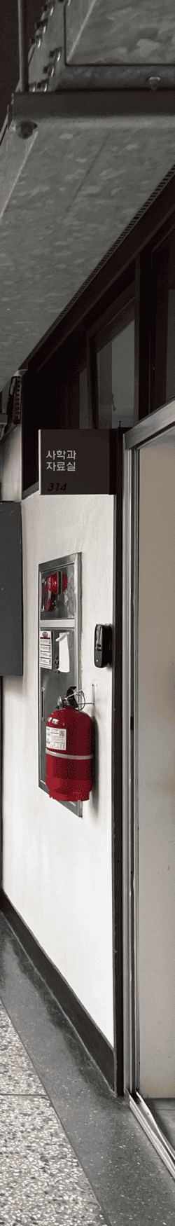 중간에 소실점이 모이는 복도를 세로로 4분할 했을 때의 왼쪽에서 네번째 사진. 오른쪽 콘크리트 벽의 위쪽에는 가로로 긴 직사각형의 나무 몰딩 창문이 있고 그 아래 사학과 자료실 안내판과 입구가 있다. 옆에는 소화기와 옥내 소화전이 있다.
