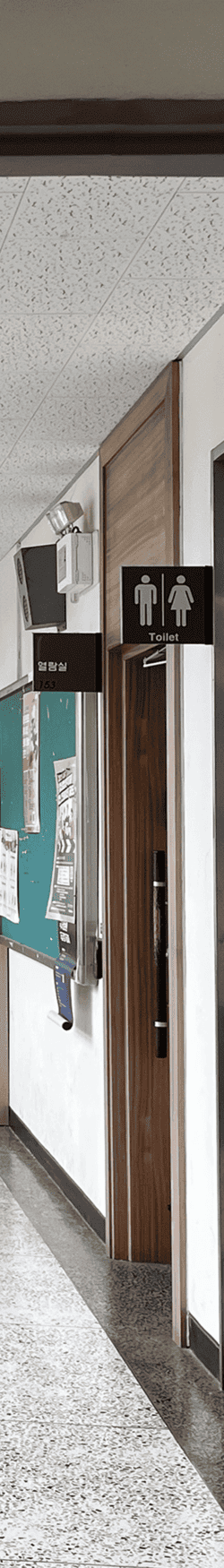 중간에 소실점이 모이는 복도를 세로로 4분할 했을 때의 왼쪽에서 네번째 사진. 콘크리트 벽에 화장실 안내판과 나무몰딩 문틀과 열람실 안내판, 가로로 긴 게시판과 그 위에 스피커가 있다. 천장은 흡음 택스 천장.