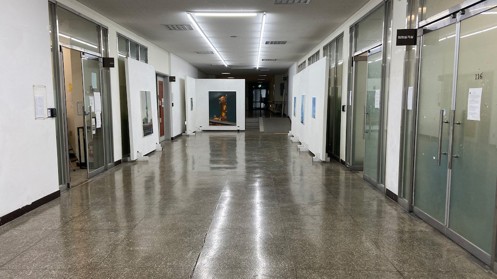 영남대학교 미술관 복도 이미지. 앞으로 길게 복도가 이어져있다. 양쪽 옆에 강의실로 들어갈 수 있는 불투명한 유리문이 있다.