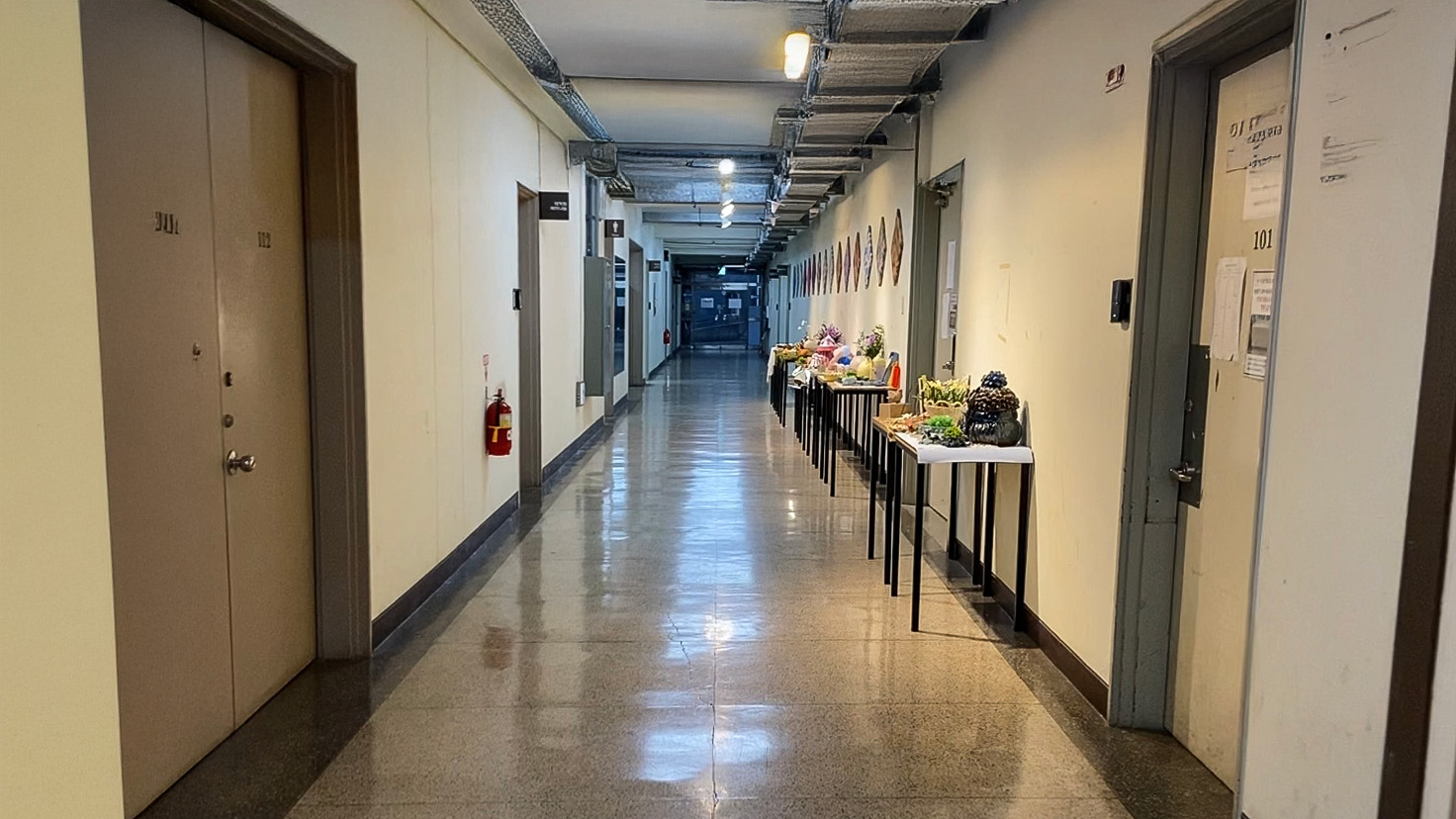 영남대학교 디자인관 복도 이미지. 앞으로 복도가 이어져있다. 노란색 조명이 켜져있다. 복도 끝에는 바깥으로 이어지는 투명한 유리문이 달려있다.왼쪽에는 가로로 놓여진 나무 책상들 위에 조형물들이 올려져있다. 양 옆으로 철제문이 있다