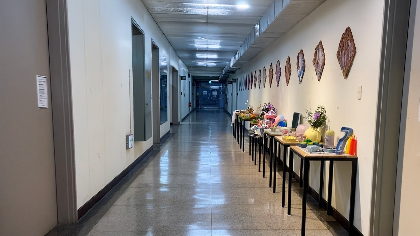 영남대학교 디자인관 복도 이미지. 앞으로 복도가 이어져있다. 복도 끝에는 바깥으로 이어지는 투명한 유리문이 달려있다. 노란색 조명이 켜져있다. 왼쪽에는 가로로 놓여진 나무 책상들 위에 조형물들이 올려져있다. 양 옆으로 철제문이 있다