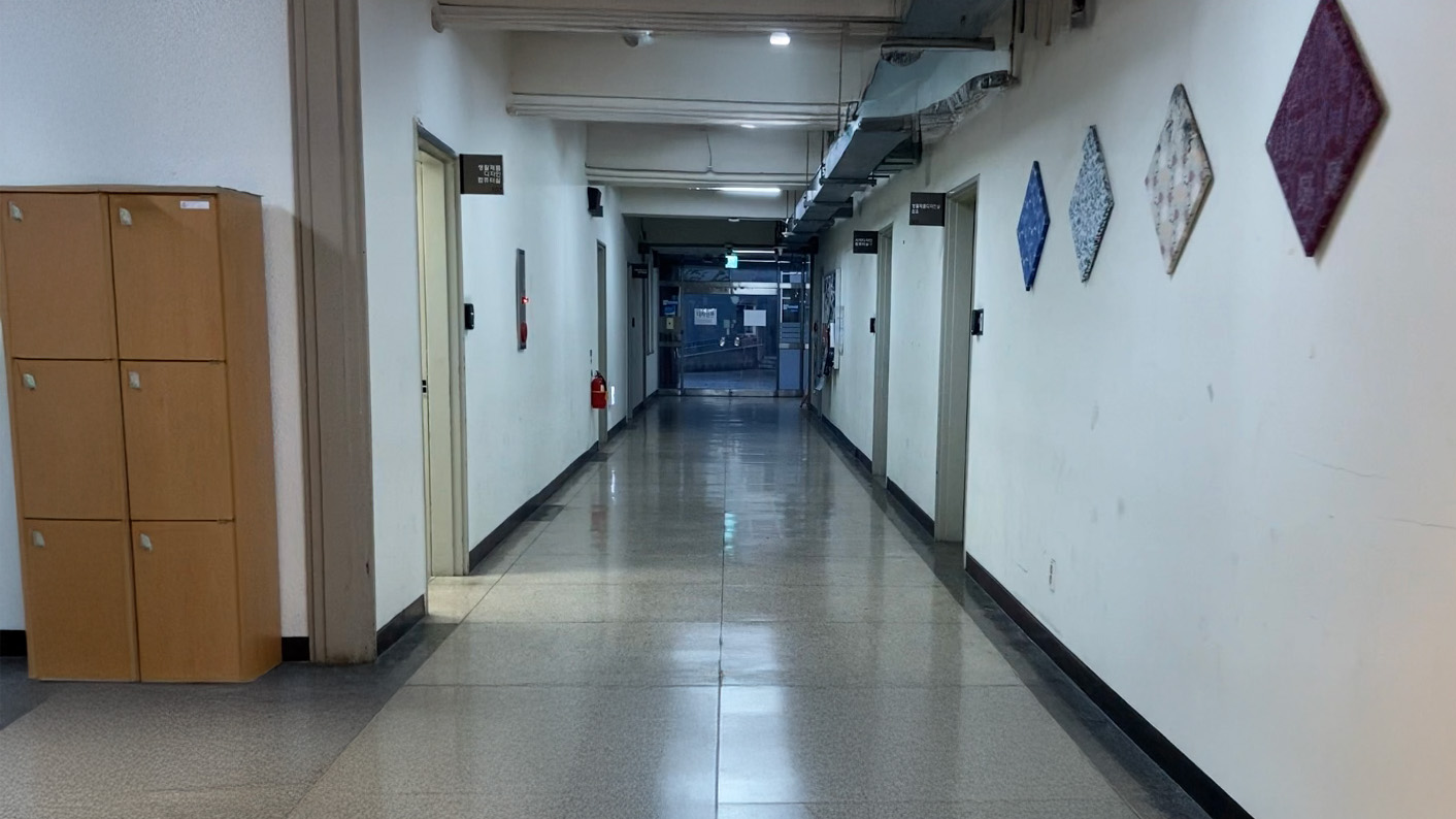 영남대학교 디자인관 복도 이미지. 앞으로 복도가 이어져있다. 복도 끝에는 바깥으로 이어지는 투명한 유리문이 달려있다. 흰색 조명이 켜져있다. 오른쪽 벽에는 마름모 형태의 캔버스가 걸려져있다. 왼쪽에는 세로로 긴 6칸의 나무 사물함이 있다. 양 옆으로 철제문이 있다