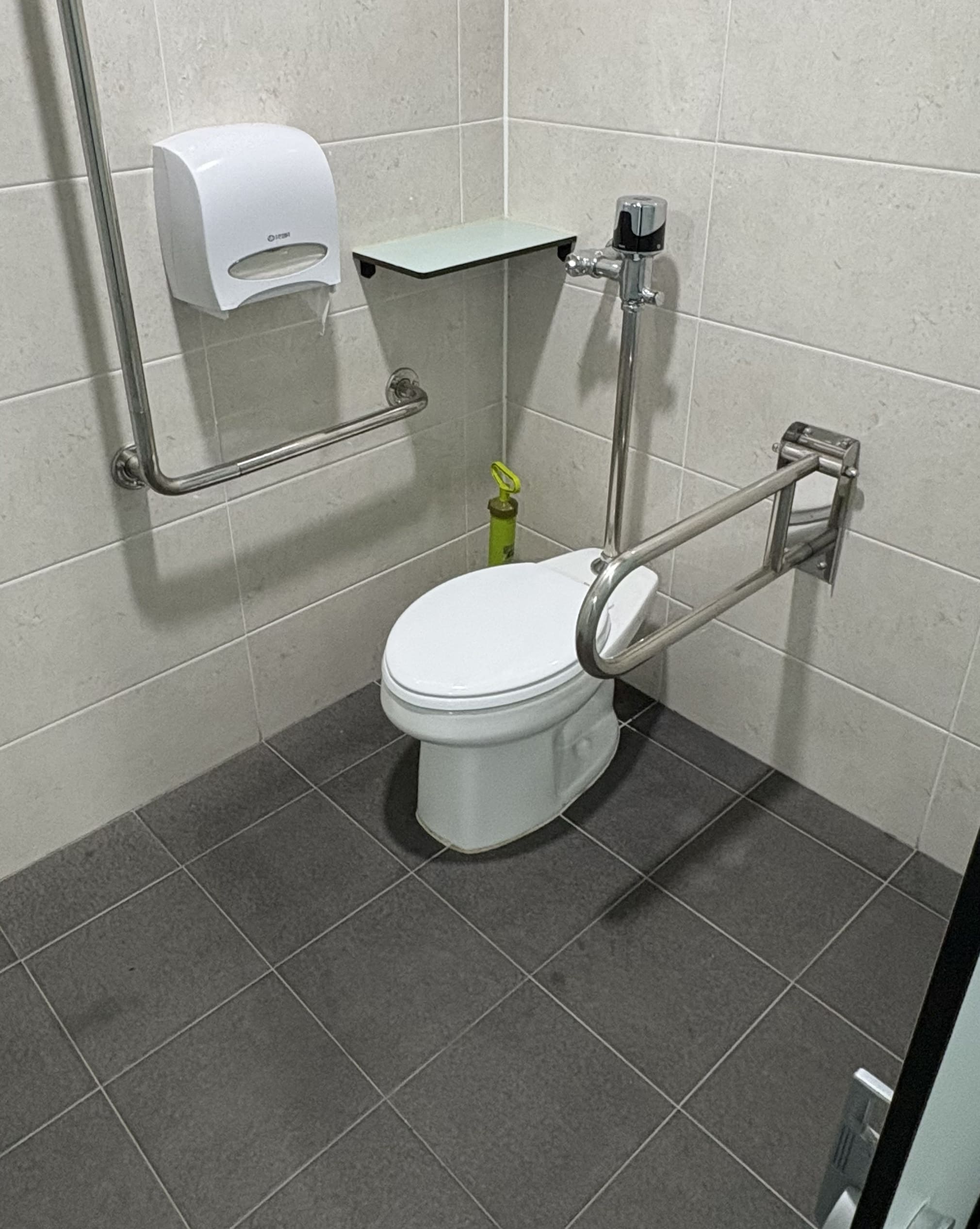 벽에는 하얀 타일 바닥에는 검은 타일 벽에는 ㄴ모양 쇠봉과 하얀 휴지통이 붙어있다. 양변기가 중간에 있고 그옆에는 둥근 손잡이가 있다.