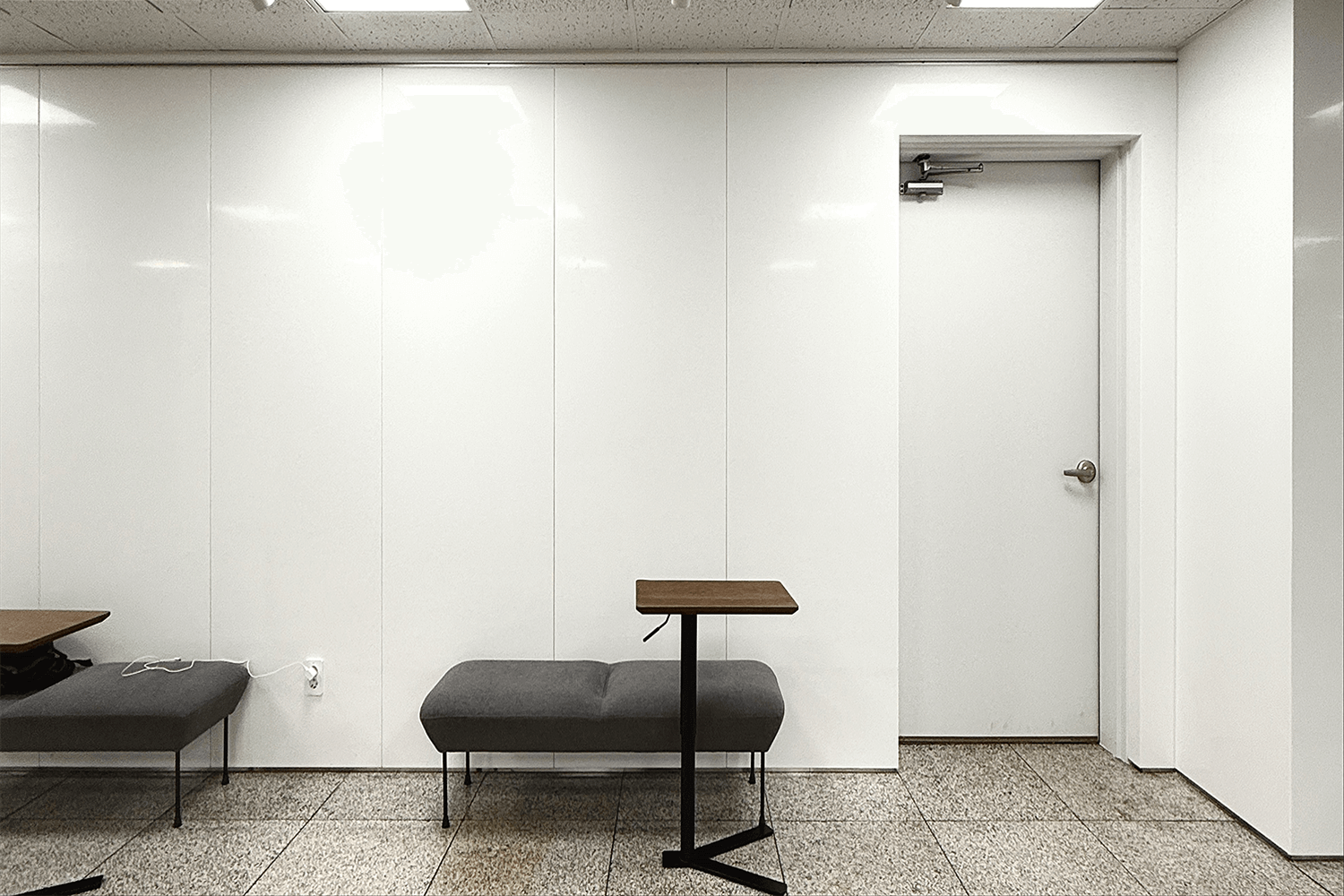 하얀색 벽이 있으며, 한 쌍의 회색 의자와 작은 나무 책상들이 연속적으로 있는 모습이다. 헉샹들을 위한 실내 대기 공간처럼 보인다.