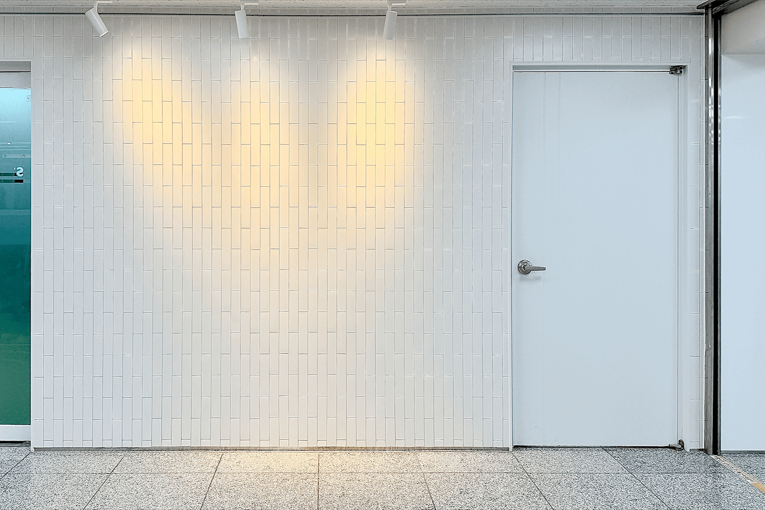 사진 전체 면적의 90%가 세로로 된 타일 형식의 벽으로 보인다. 흰색 벽에는 노란빛의 조명이 비치고 있으며 사진의 오른쪽에는 흰색 문이 위치하고 있다.