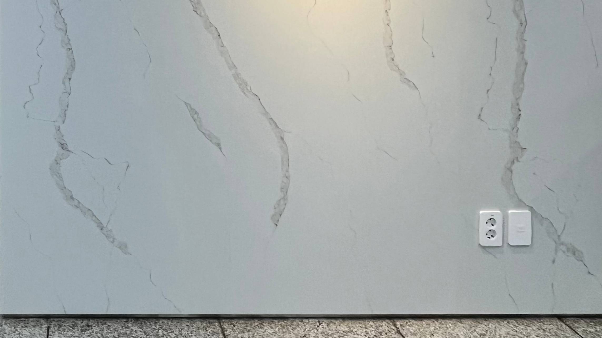 밝은 회색 대리석 벽 사진. 전체는 매끈한데, 여기저기 연한 회색 줄무늬가 자연스럽게 흐르듯 퍼져 있음. 바닥은 까슬까슬한 화강암 타일이다.