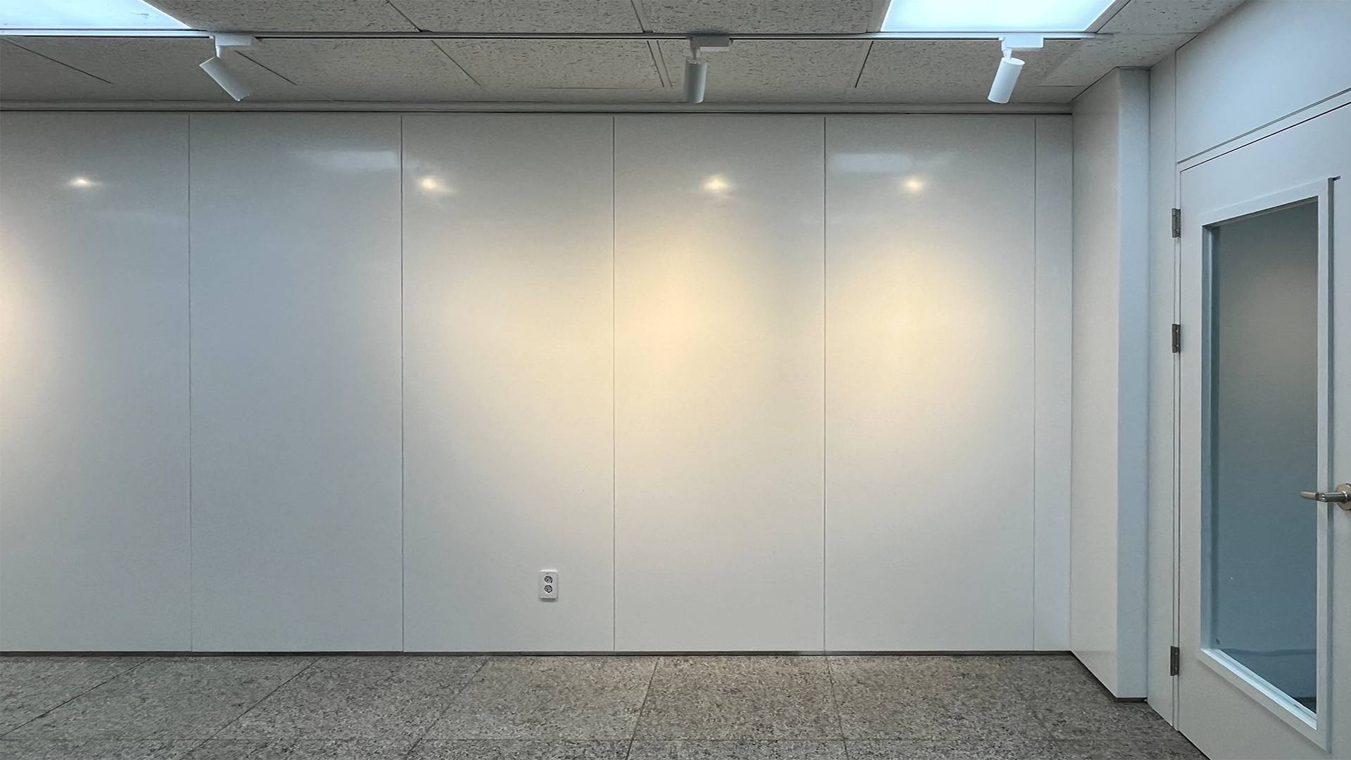 깨끗한 흰색의 반들반들하고 매끈한 벽 사진. 수직 방향으로 얇은 선이 여러 개 나있어서 넓은 벽이 여러 개로 나뉘어있다. 각각의 판넬 위에는 조명이 비춰져 빛이 동그랗게 번져 있다. 가운데쯤에는 하얀 콘센트가 붙어 있다