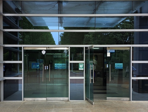 한 유리벽이 5개의 흰 창틀로 인해 가로로 나뉘며, 정중앙에서 양쪽으로 열리는 구조의 투명한 유리문 두 짝이 나란히 설치되어 있다.