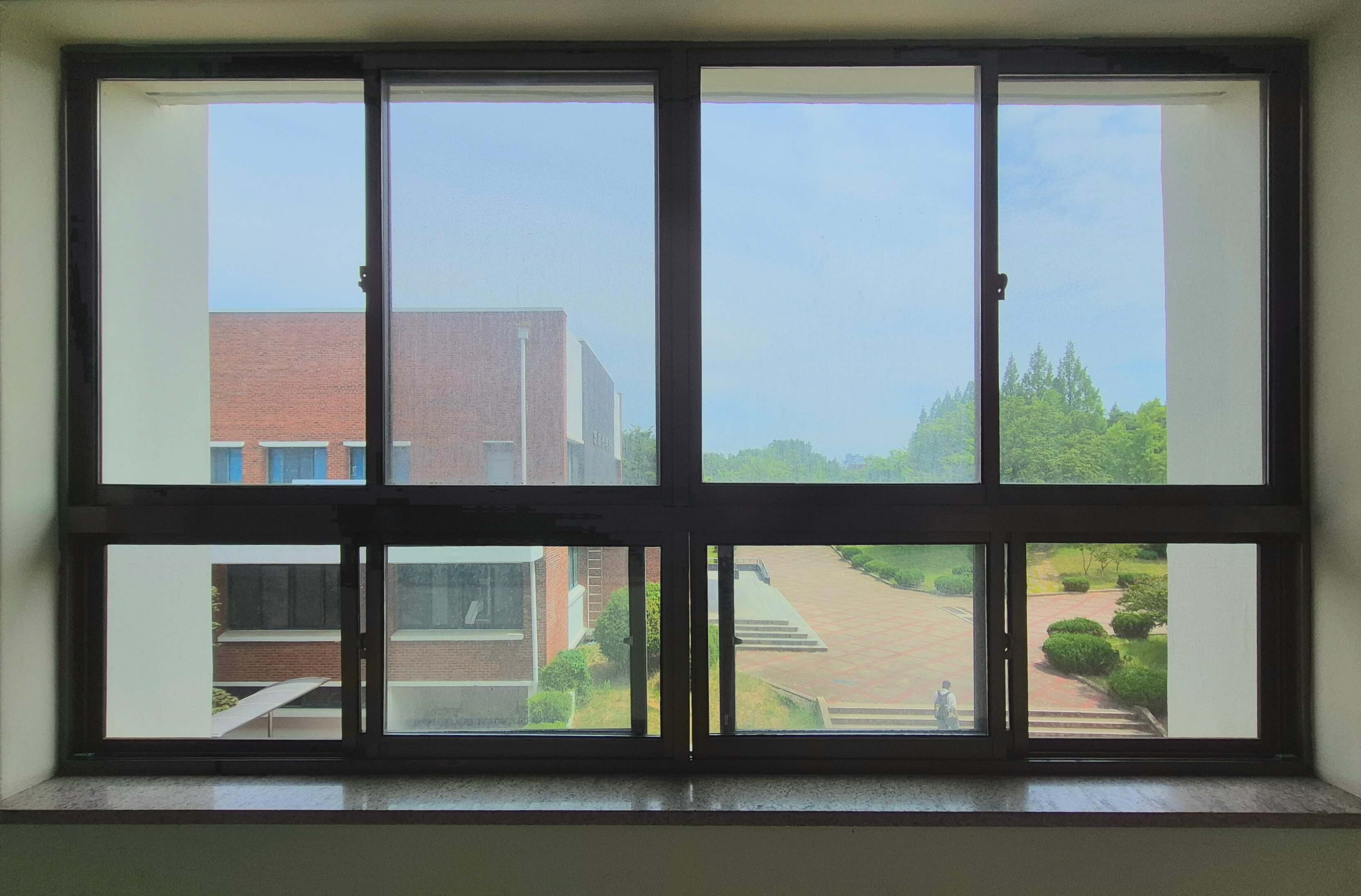 인문관 창문, 흰 벽 위로 검은 창틀로 된 직사각형 형태의 창문이다. 세로로 긴 직사각형 네 개가 위쪽에 있고, 그 아래쪽에 가로로 긴 직사각형 네 개가 나란히 붙어있는 형태이다. 창문 너머로 붉은 벽돌벽 견물과 나무가 보인다.