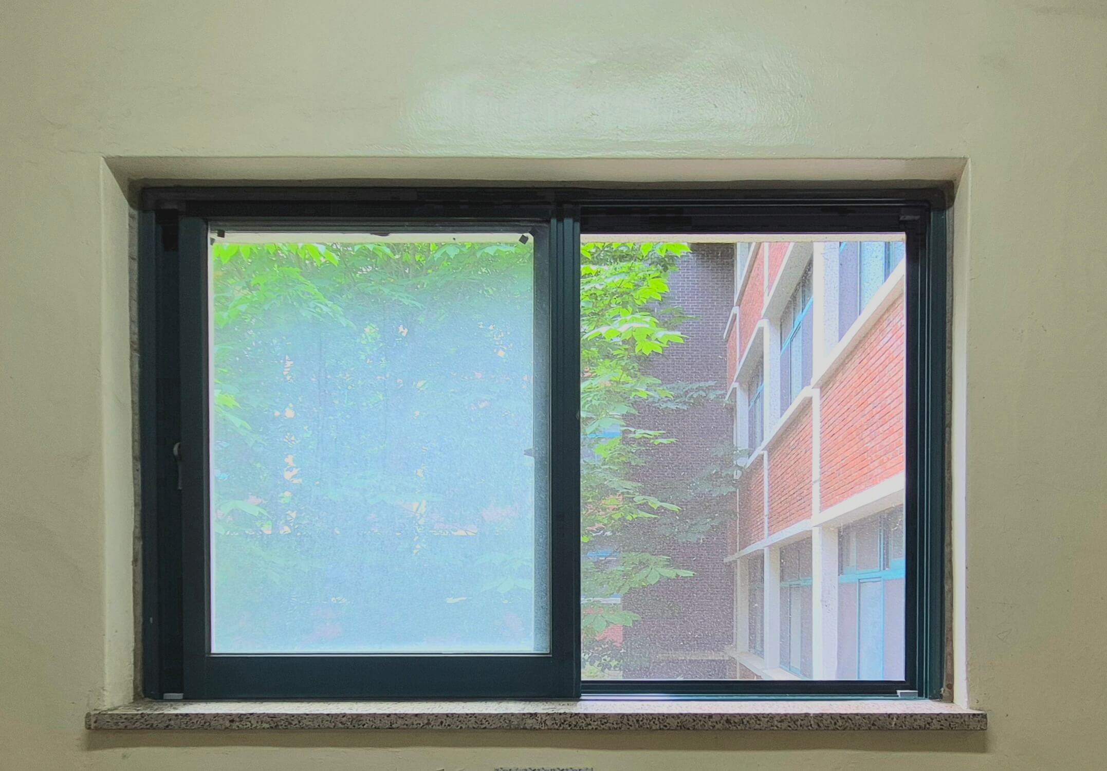 인문관 창문, 흰 벽 위로 검은 창틀로 된 직사각형 형태의 창문이다. 두 개의 창문 중 왼쪽의 창문은 습기가 차서 바깥이 잘 보이지 않는다. 오른쪽 창문 너머로는 왼쪽으로 나무가, 오른쪽으로 묽은 벽돌벽 건물의 일부가 보인다.